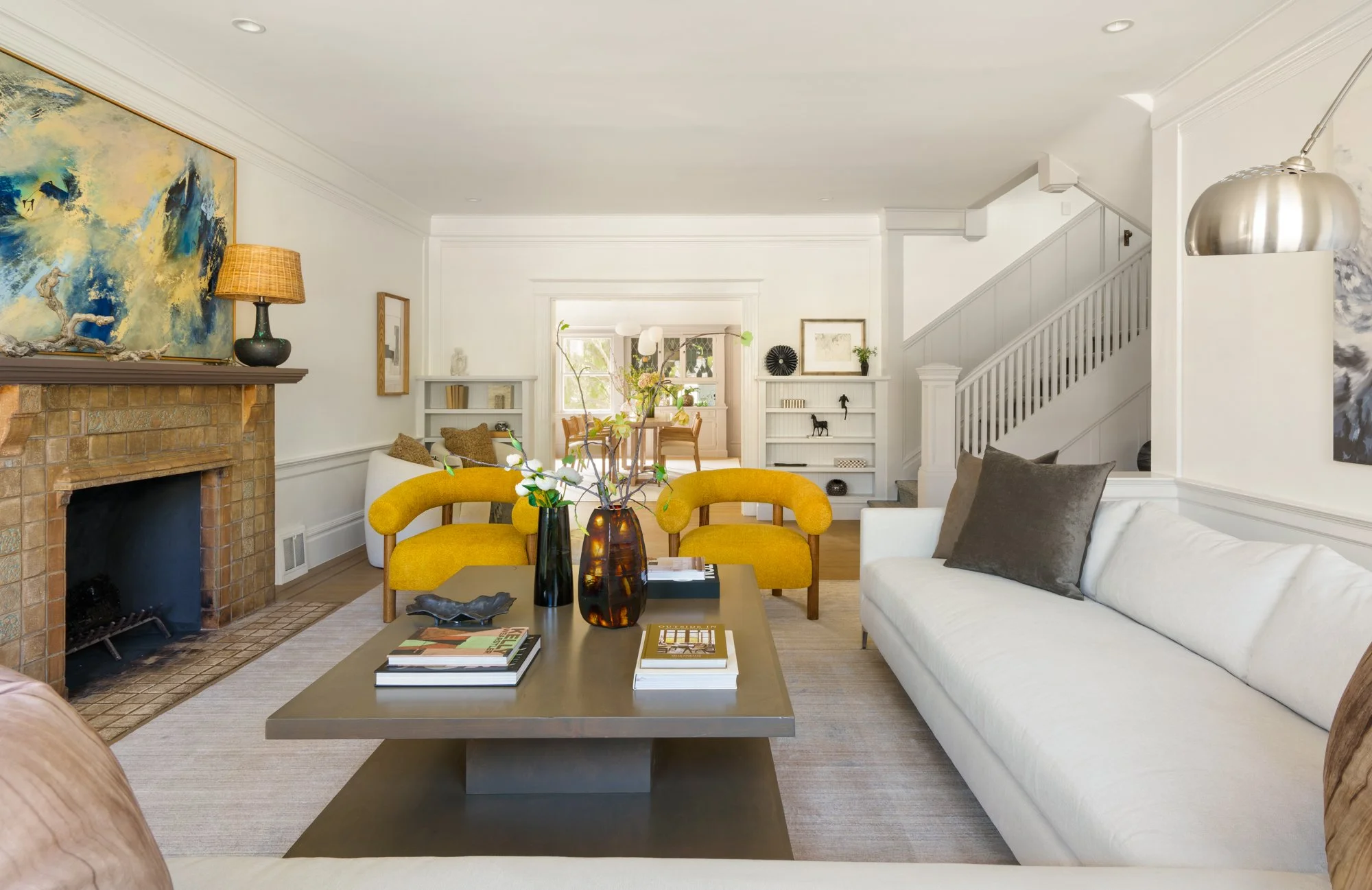 Spacious living room with modern decor, featuring yellow armchairs, a white sofa, a large coffee table, abstract art above a fireplace, and bookshelves, leading to a dining area with a staircase on the right.