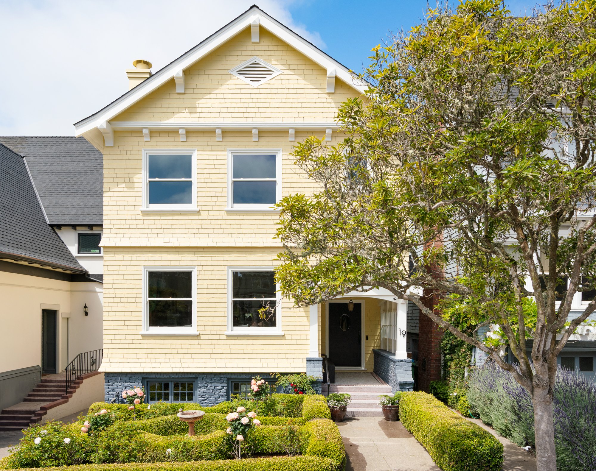 Yellow two-story house with a gabled roof, surrounded by a manicured garden with hedges and flowers, and a large tree in the front yard.
