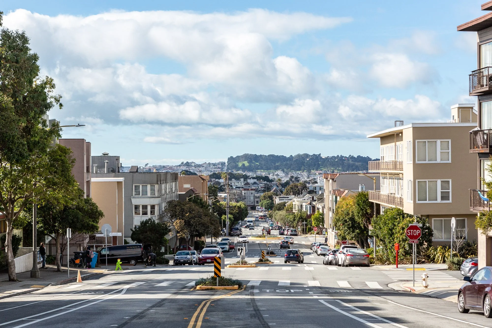Urban street with buildings, cars, and trees under a blue sky with clouds.