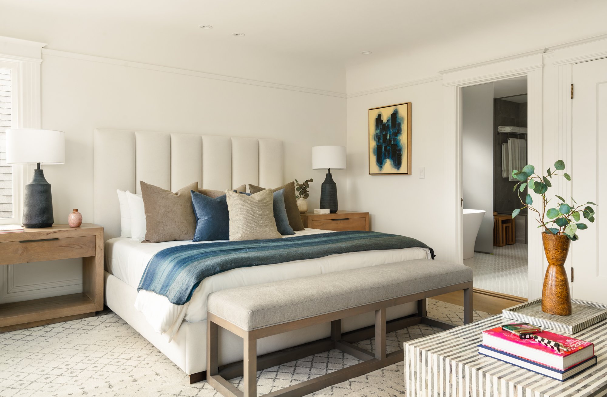 Modern bedroom interior with a large bed featuring white and blue bedding, a cushioned headboard, wooden nightstands with lamps, a bench at the foot of the bed, abstract wall art, a small table with books and a vase, and an open doorway leading to a 