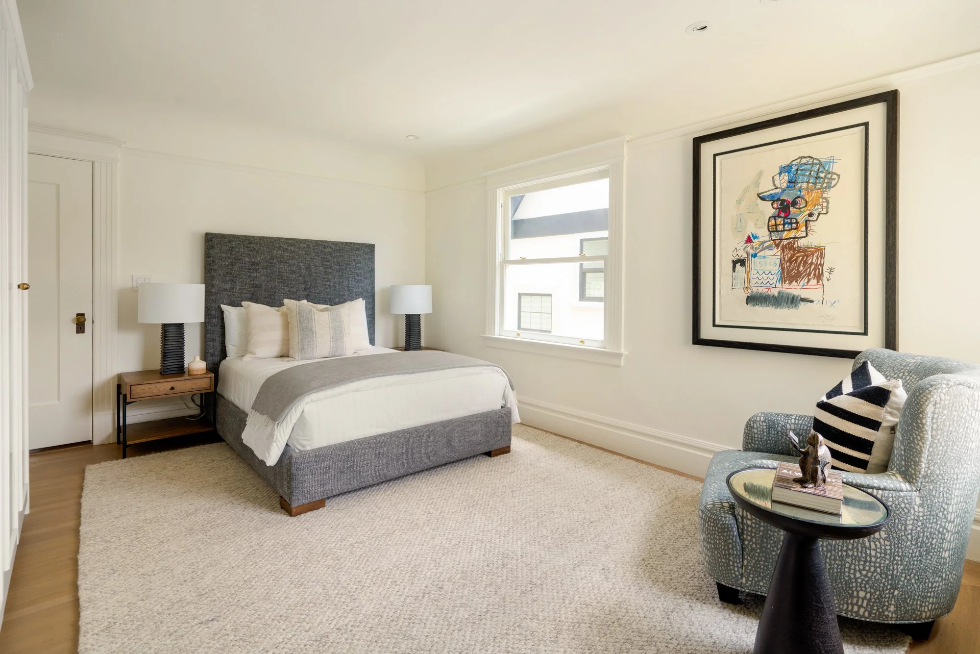Modern bedroom with a gray upholstered bed, two side tables with lamps, abstract art, a plush chair with a striped pillow, and a small round side table.
