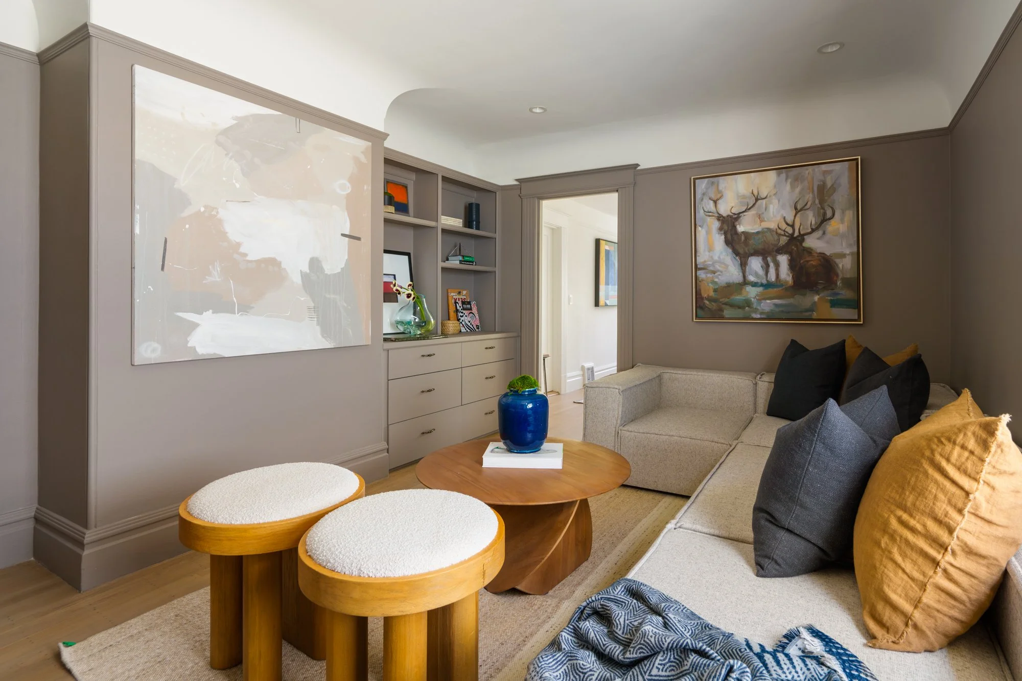 Contemporary living room with beige walls, abstract and deer artwork, built-in shelves, a beige sectional sofa, wooden stools, and a blue vase on a round wooden coffee table.