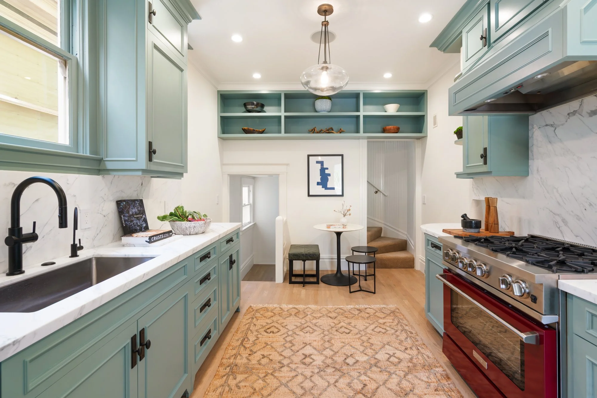 Modern kitchen with blue cabinets, marble countertops, red oven, and black faucet. A small dining area with a white round table and stairs leading up.