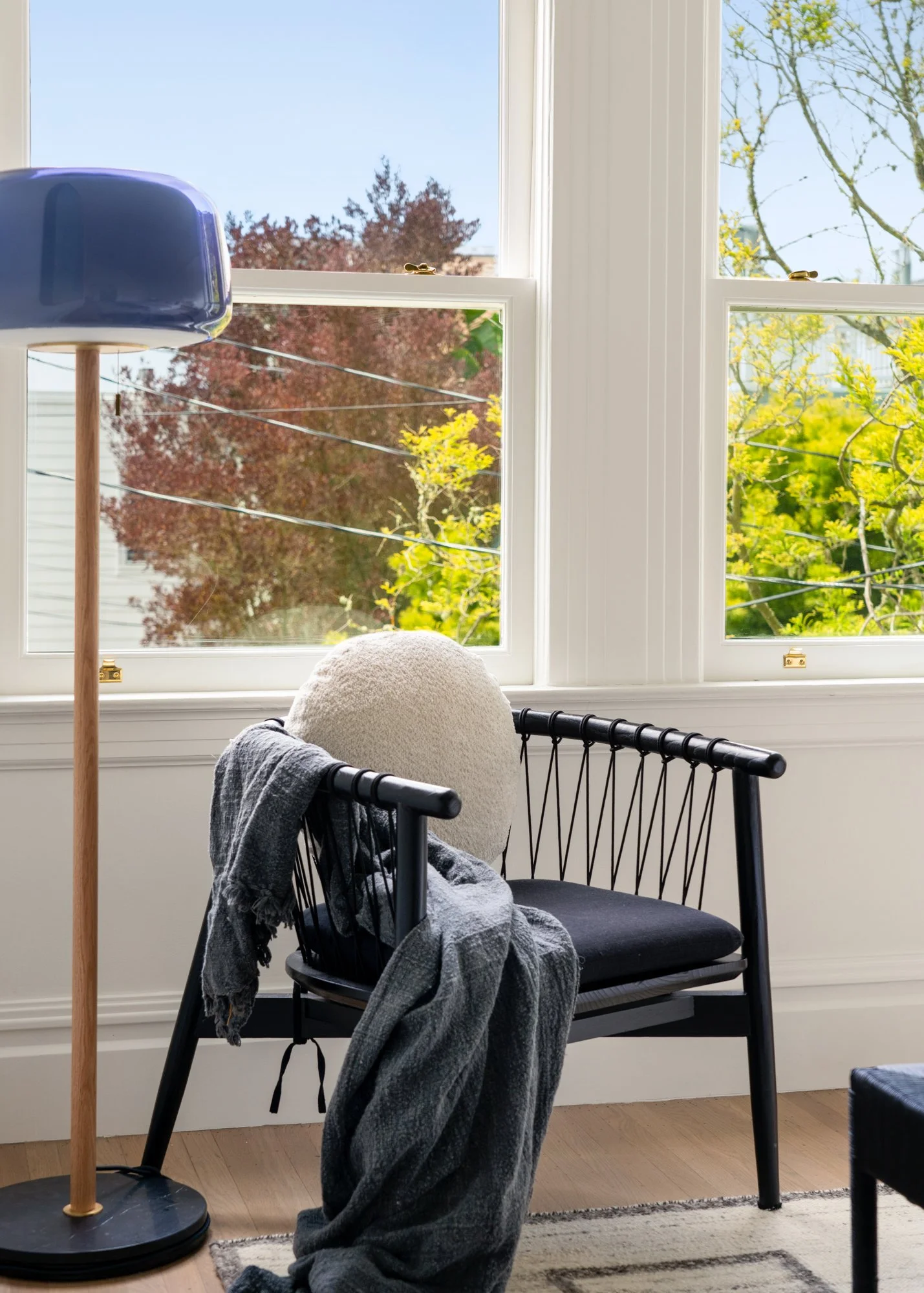 Cozy living room corner with black modern chair, round cushion, dark gray throw blanket, and wooden floor lamp near a window showing vibrant trees.