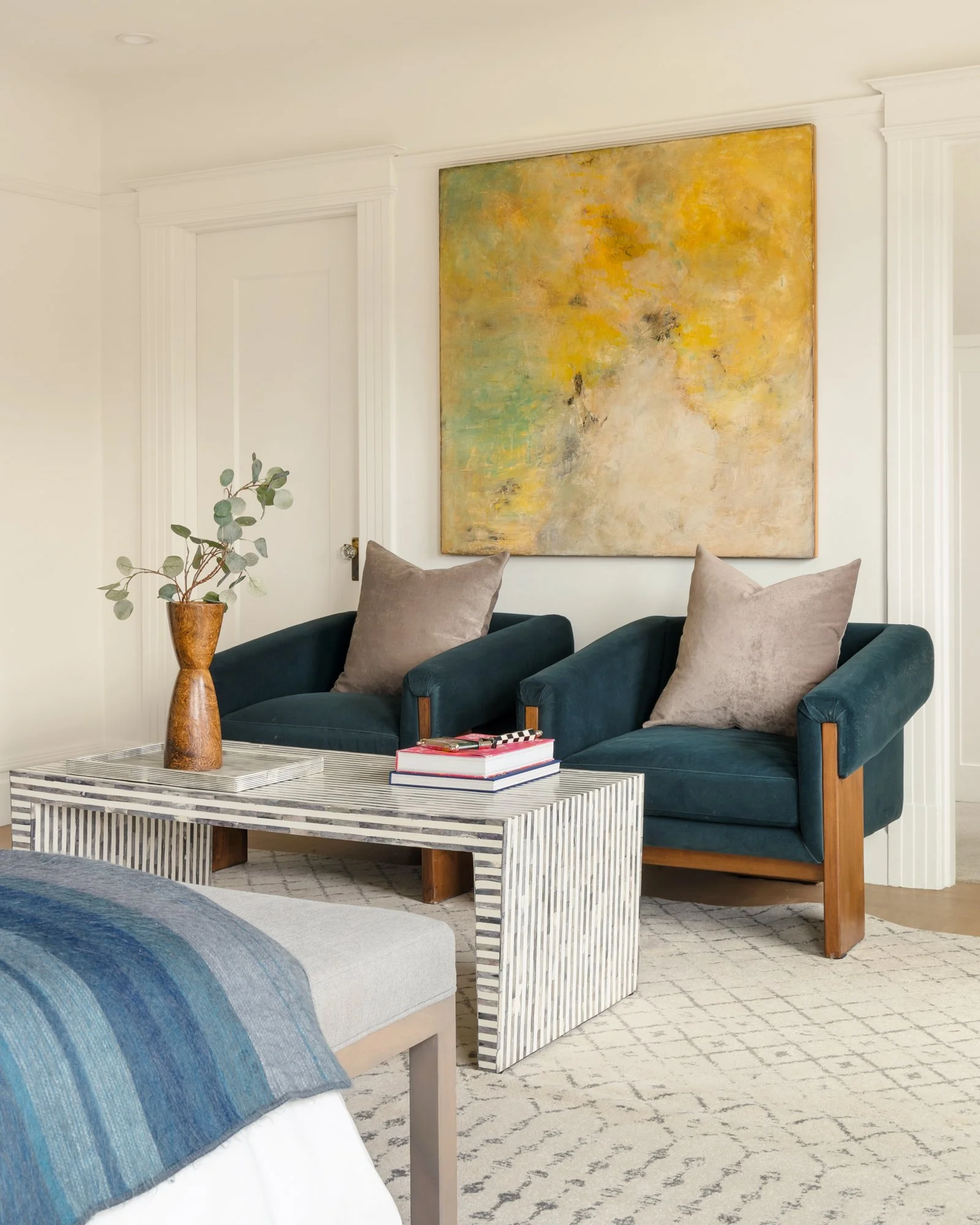 Modern living room with two teal armchairs, beige pillows, striped coffee table, wooden vase with leaves, abstract yellow painting on the wall, and a blue throw on the bed.