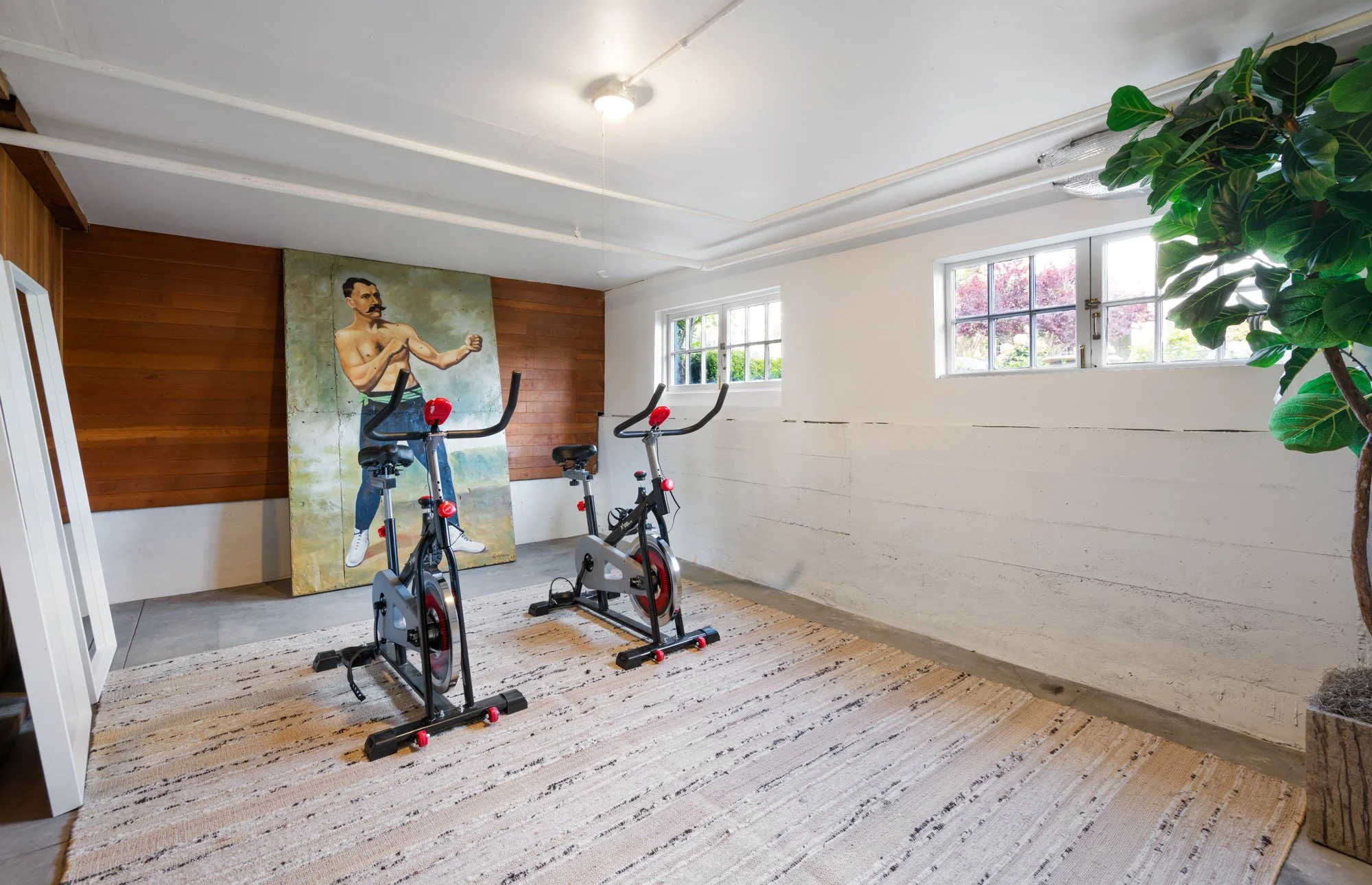 Room with two stationary bikes, large vintage boxing poster, and potted plant.