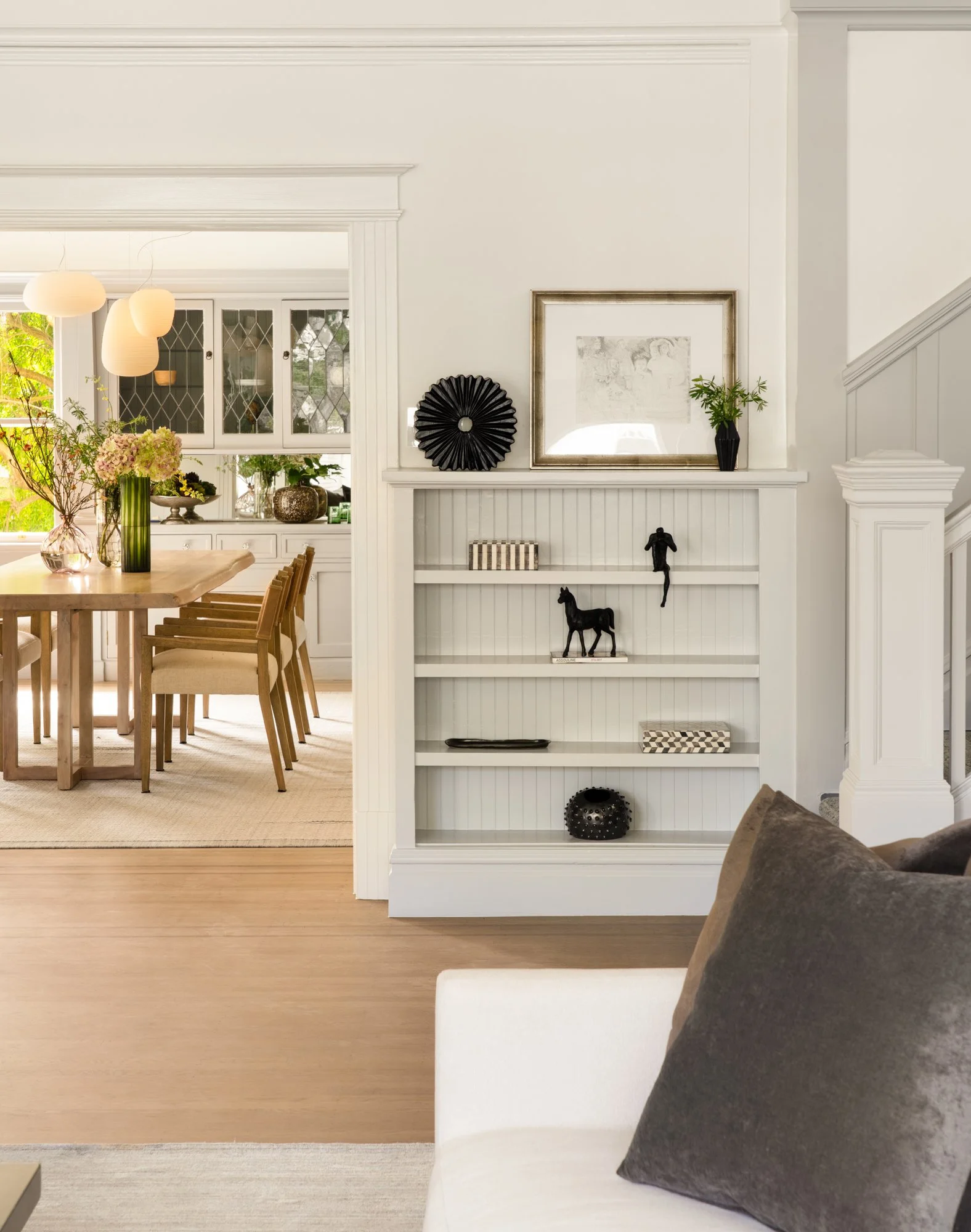 Living room with white walls and built-in shelf, featuring black sculptures and framed artwork. Adjacent dining room with wooden table and chairs, pendant lights, and decorative plants.