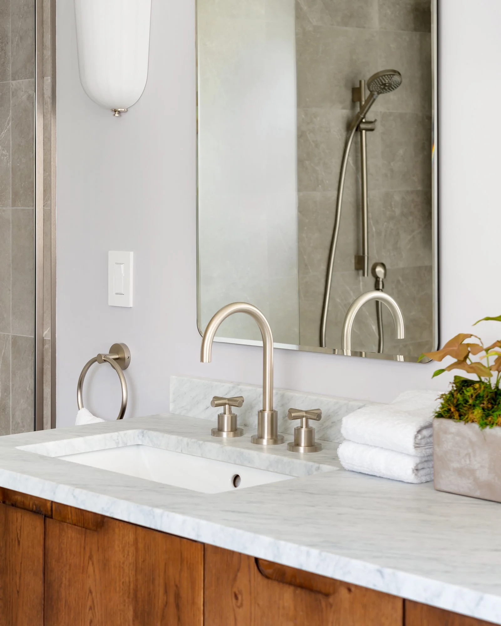Modern bathroom with a marble countertop, stainless steel faucet, mirror, and wooden cabinetry, featuring a potted plant and stacked white towels.