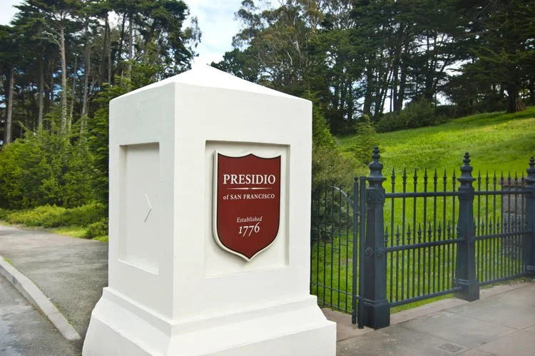 Sign at the entrance of the Presidio of San Francisco, established in 1776, with surrounding greenery and iron fence.