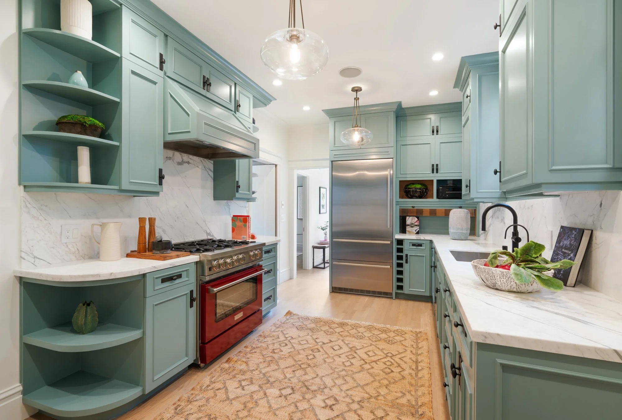 Modern kitchen with teal cabinets, white marble countertops, a stainless steel refrigerator, and a red oven. Open shelving displays decorative items. A patterned rug covers the light wood floor.