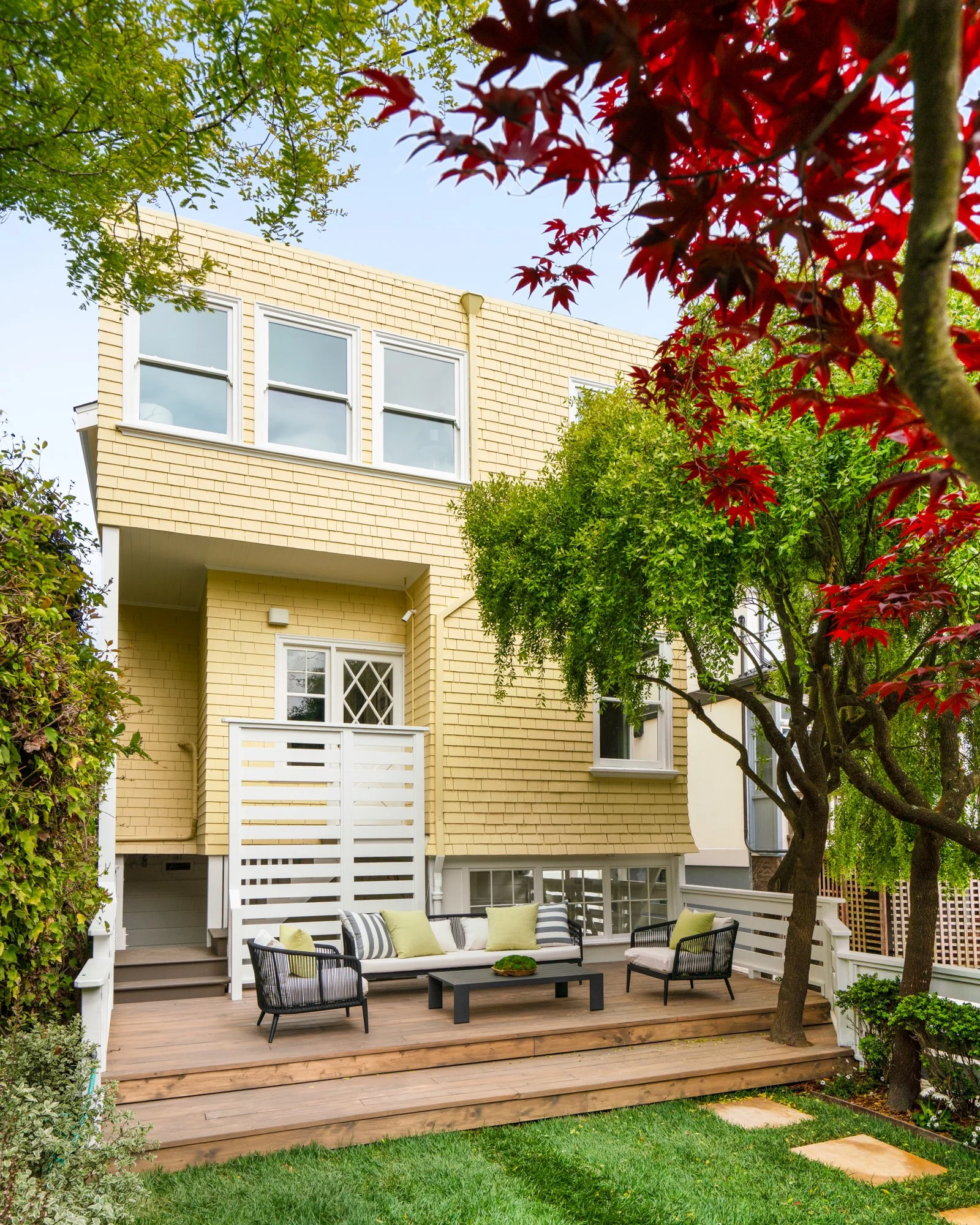 Modern backyard patio with wood deck, outdoor seating, and greenery, featuring a two-story yellow house with windows and surrounding trees.
