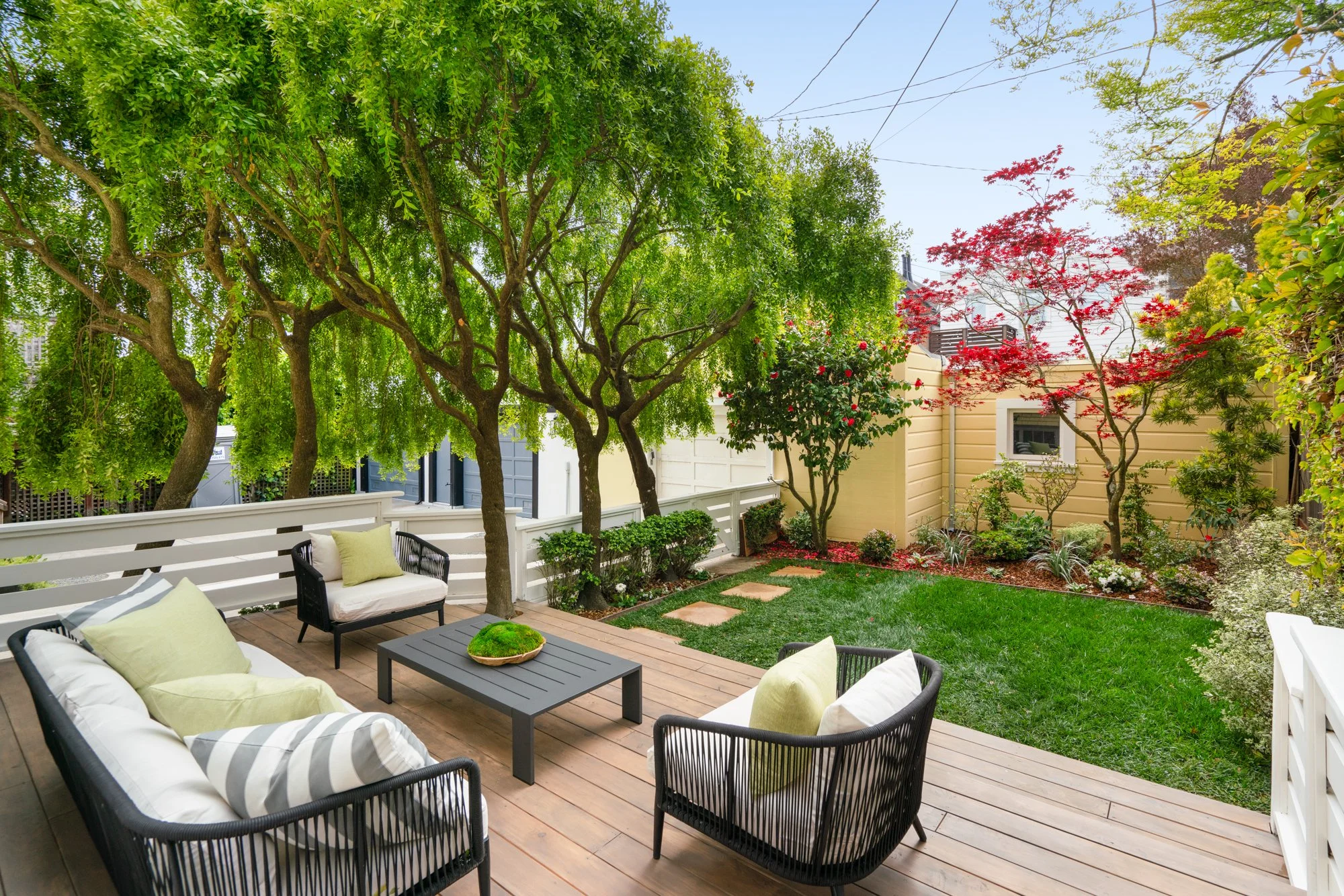 Backyard patio with wicker furniture and cushions, surrounded by greenery and trees, featuring a small lawn and garden area.