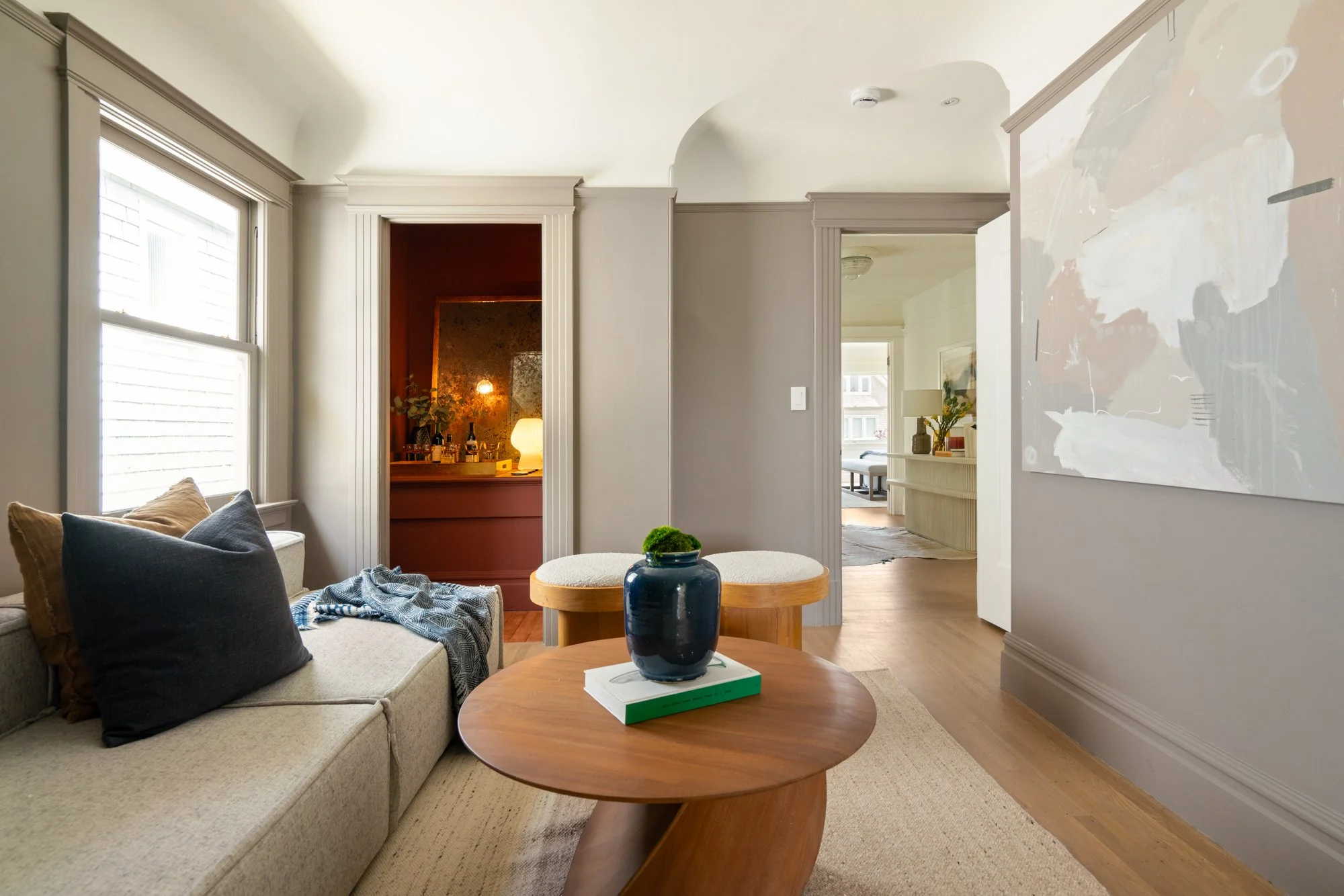 Modern living room with beige walls, a gray sofa with cushions, a wooden coffee table with a vase and books, abstract wall art, and a view into an adjoining decorated room and hallway.