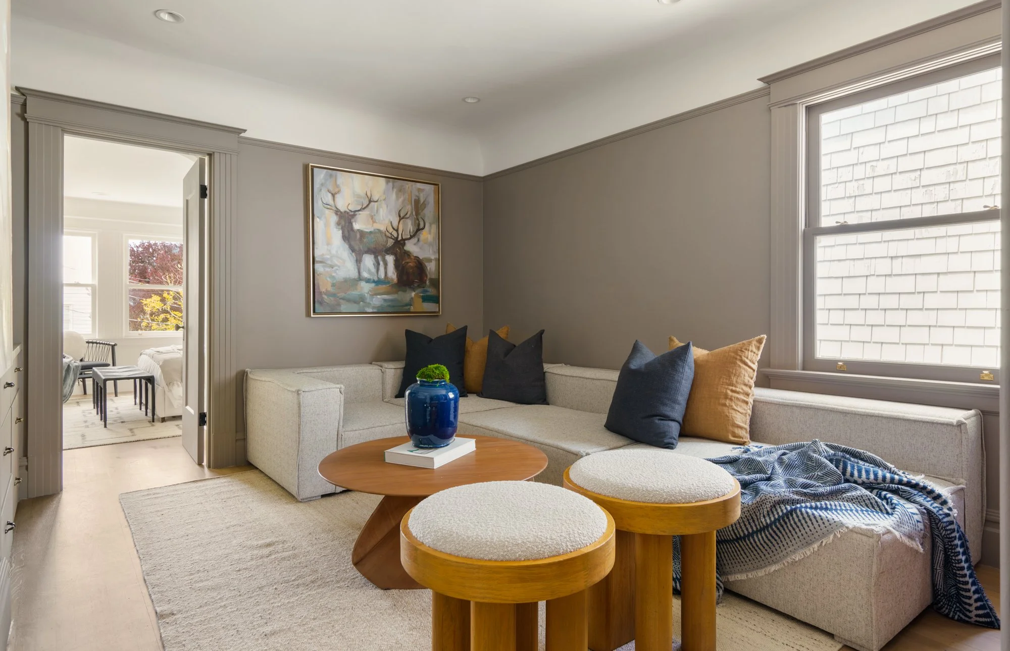 Neutral-toned living room with a corner sofa adorned with blue and brown pillows, a round wooden coffee table, and cylindrical stools. The room has beige walls and a large window. There's artwork featuring deer above the sofa, and a doorway leads to 