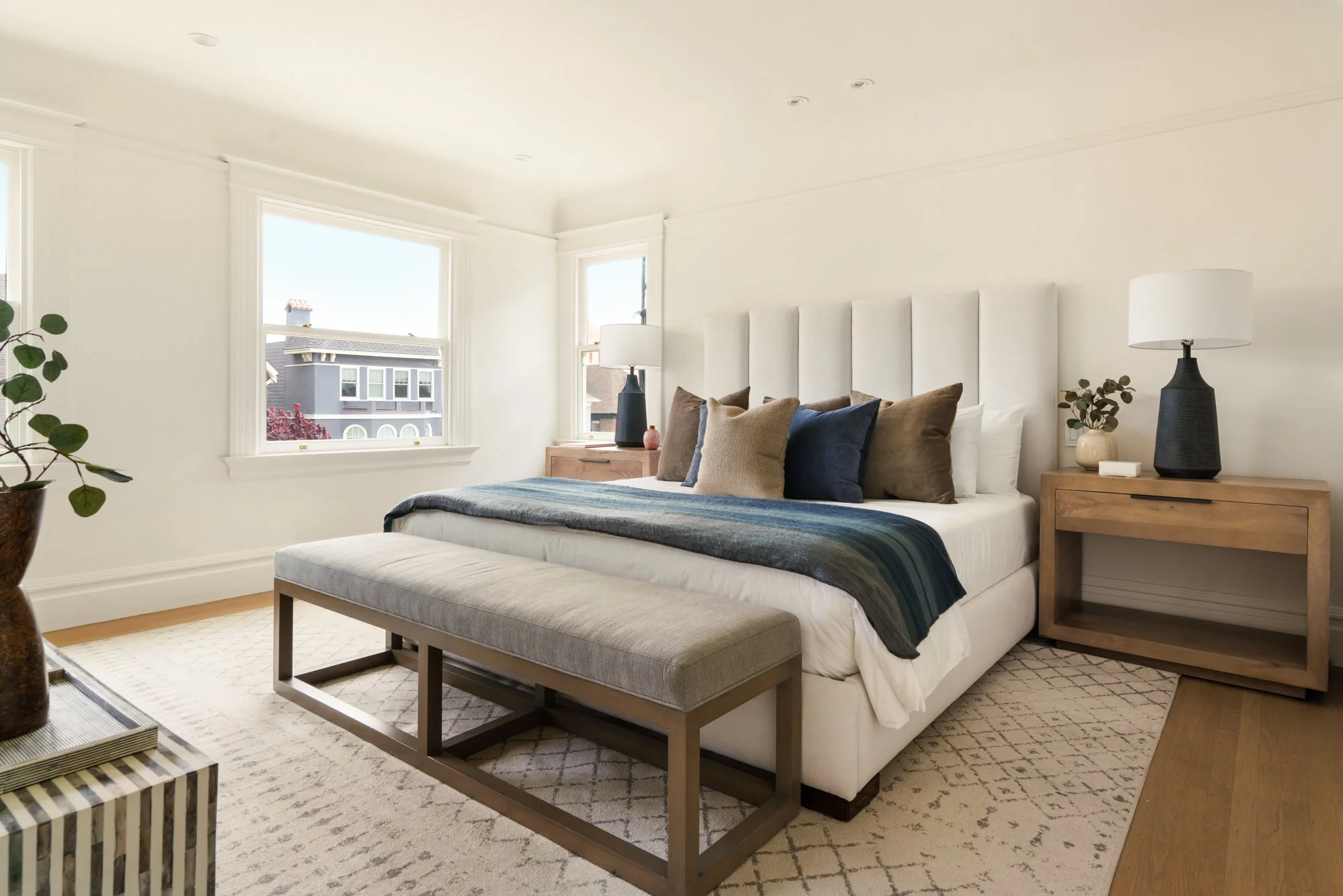 Modern bedroom with white walls, a large bed with a padded headboard, blue and brown pillows, wooden nightstands, and lamps. A bench sits at the foot of the bed, and there are large windows with natural light.