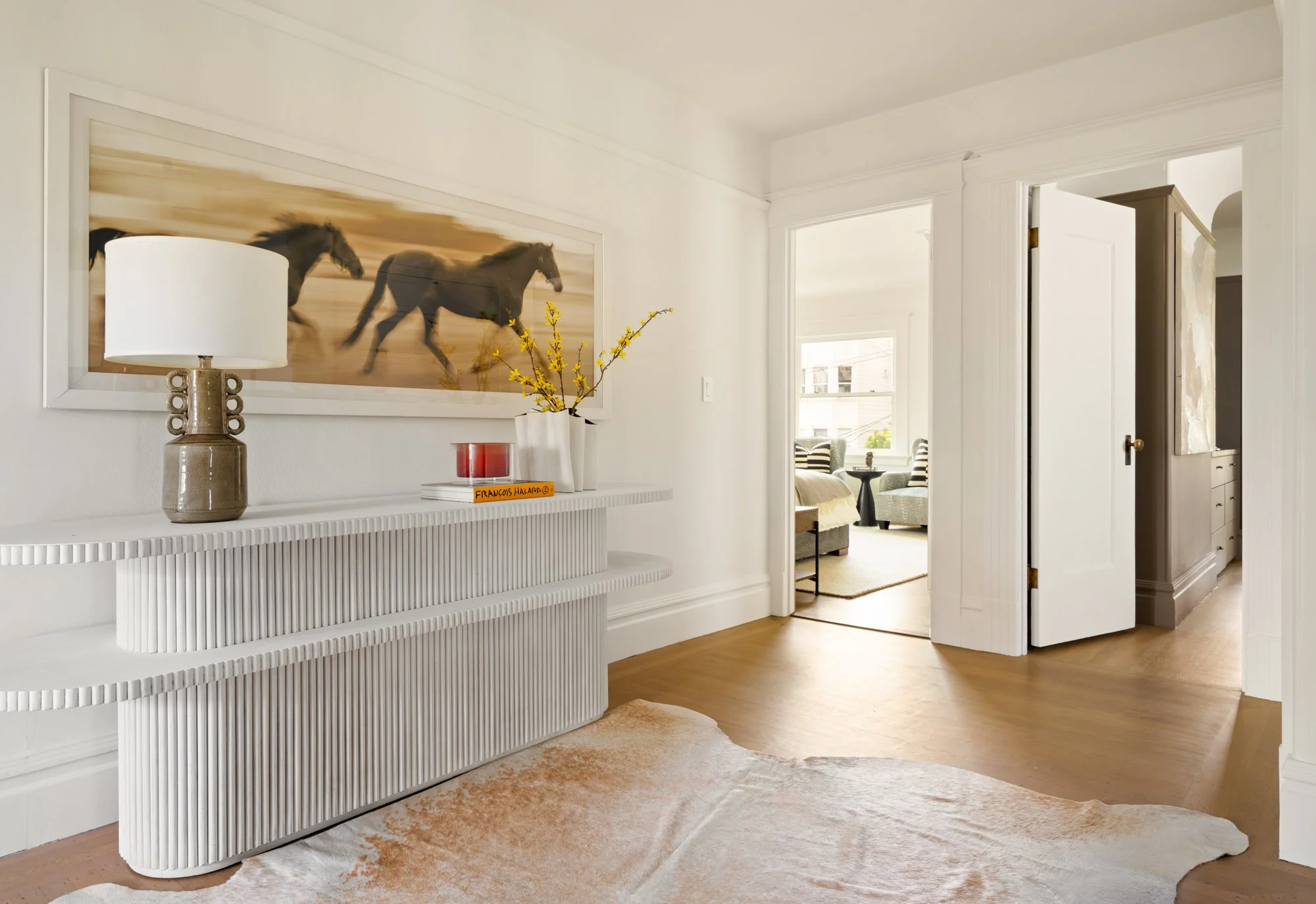 Elegant interior hallway with a white sideboard, decorative lamp, yellow flowers in a vase, and a blurred horse painting above. A doorway leads to a sunlit room with furniture in the background. Wooden floor with a cowhide rug.