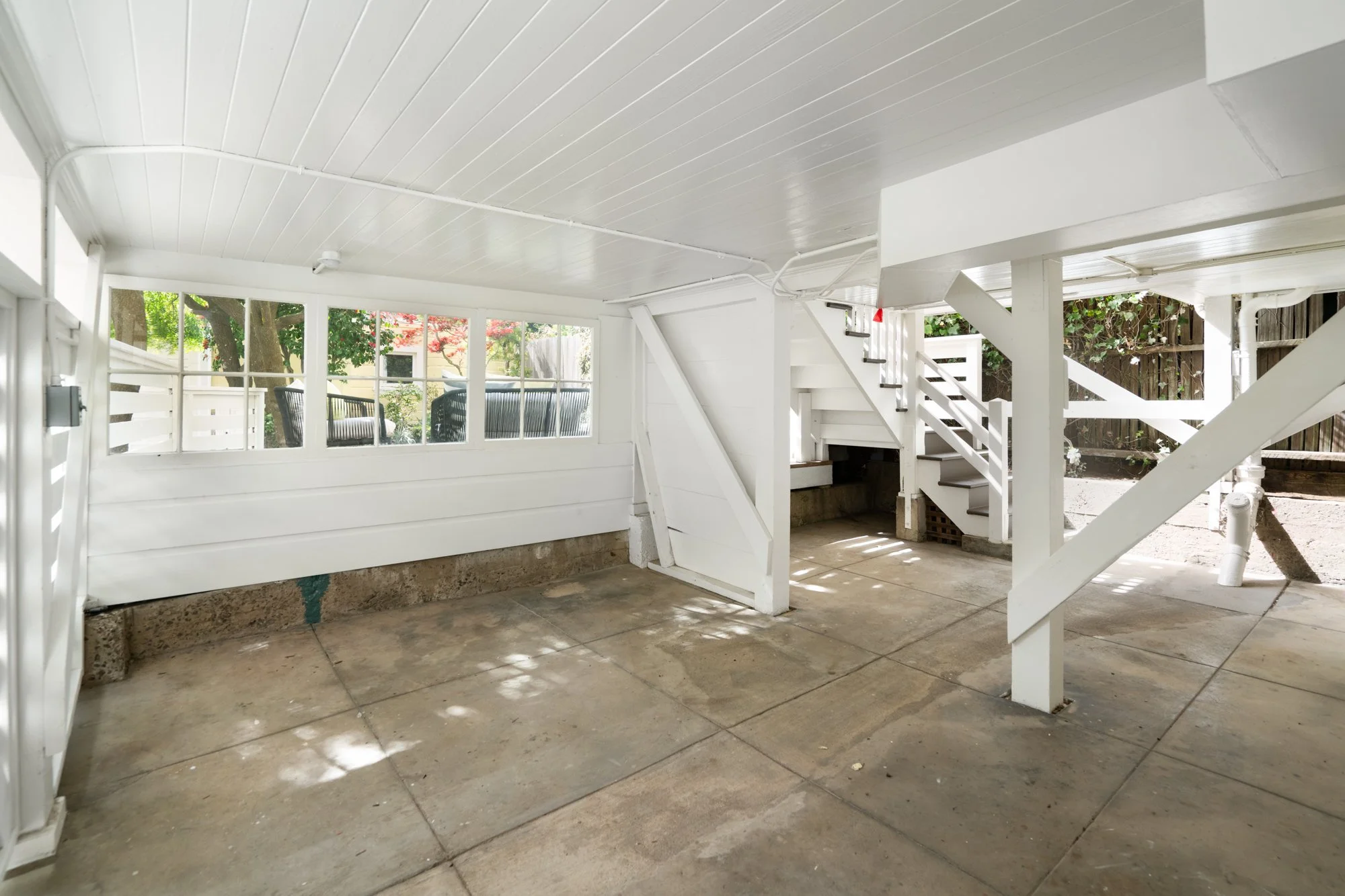 Empty white-painted outdoor carport with concrete floor, staircase, and open windows.