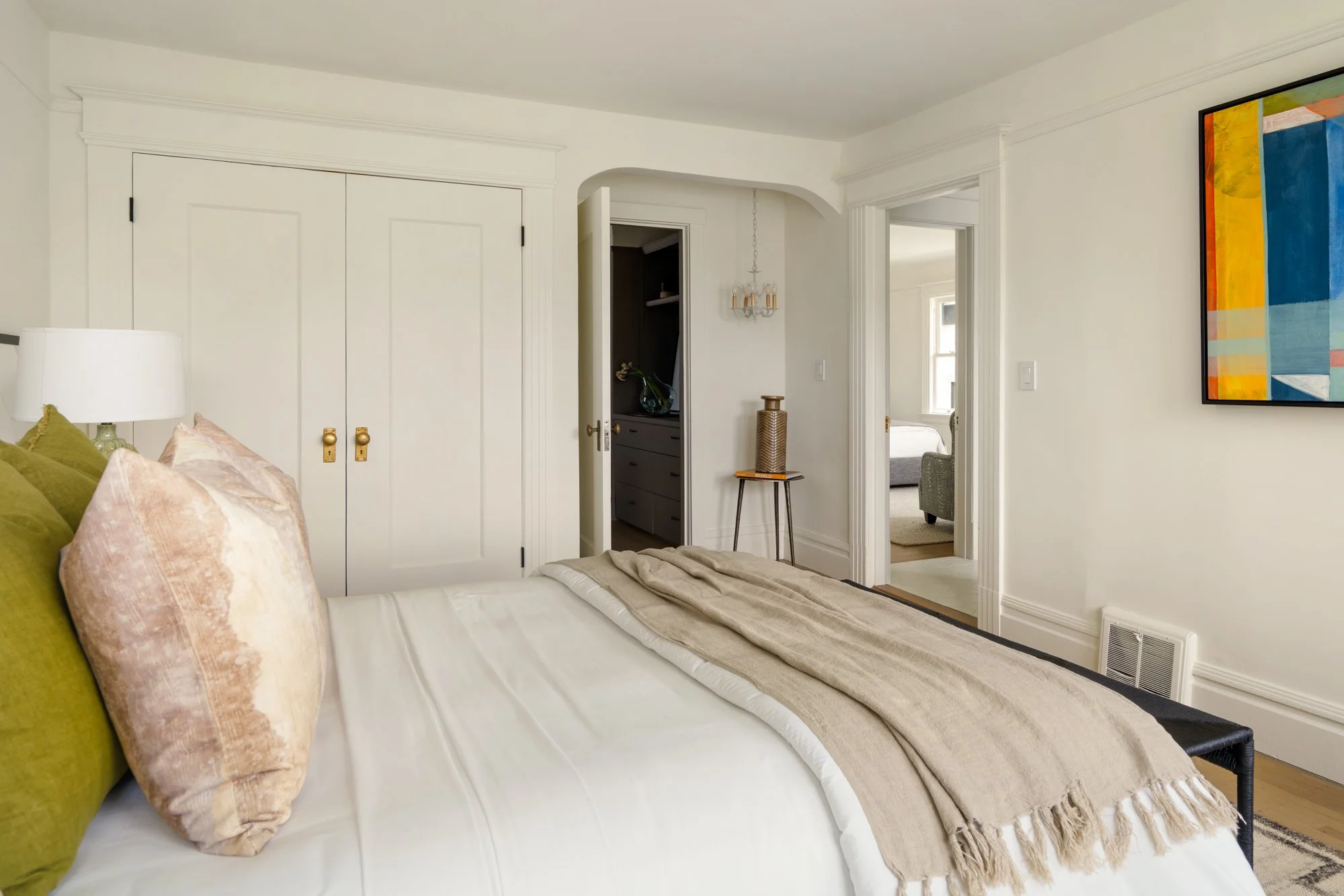Bright bedroom with white bedding and green throw pillows, featuring a closet with double doors, a modern lamp, a small round table, and colorful modern artwork on the wall.