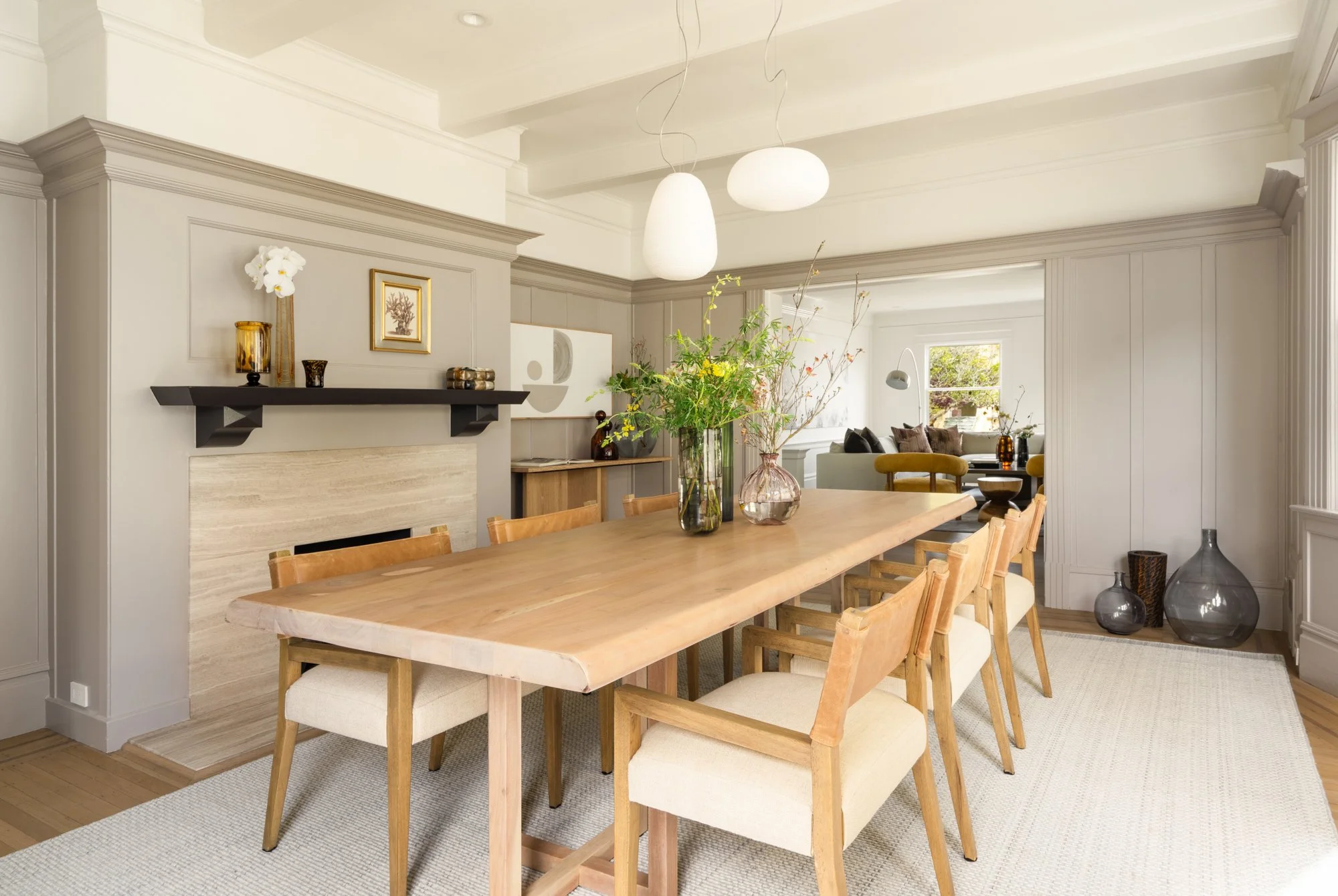 Modern dining room with a wooden table, beige chairs, and floral vases, adjacent to a living area with neutral decor and large windows.