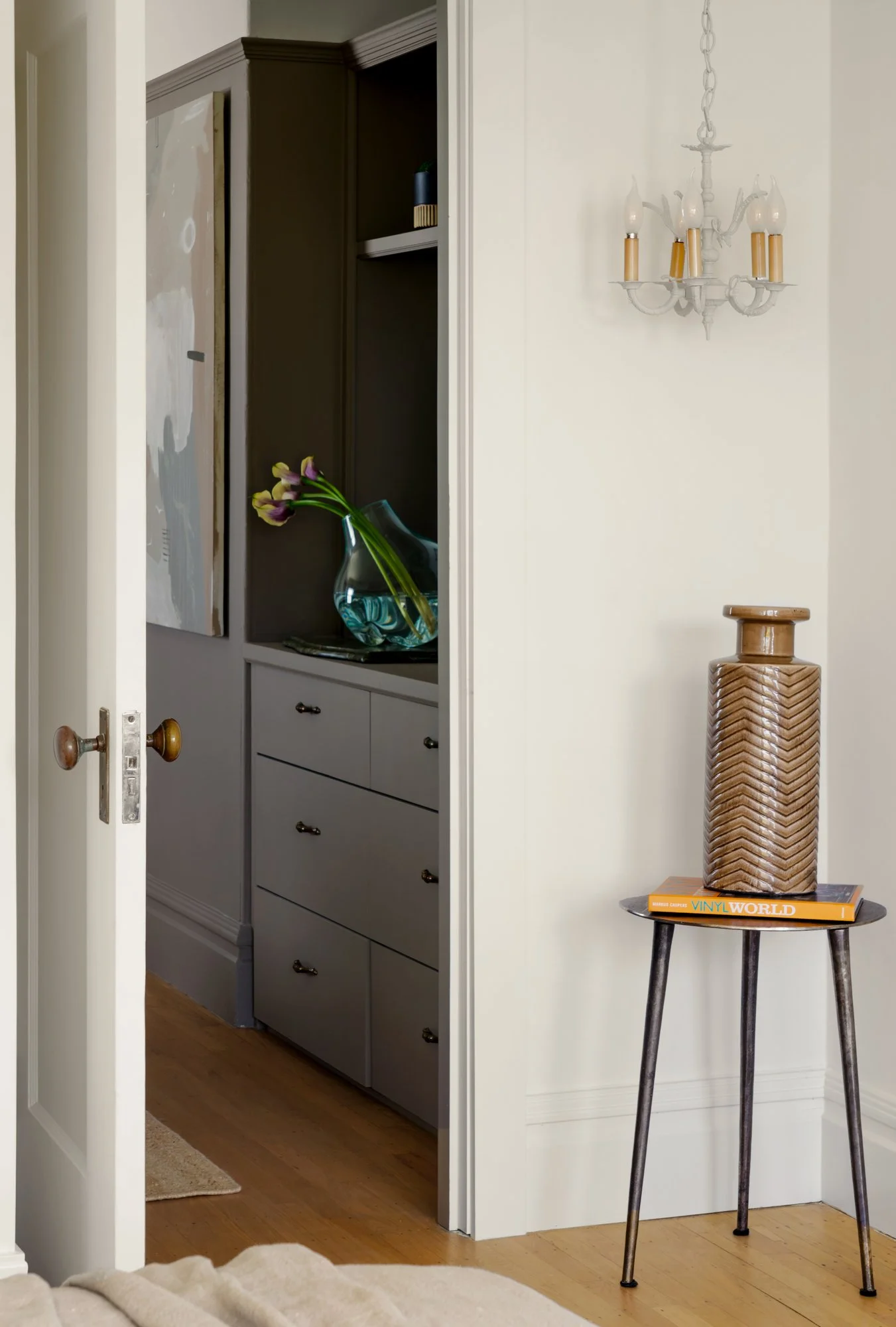 Interior featuring an open door revealing a dresser with flowers in a vase, a modern chandelier on the wall, and a small table with a decorative vase and a book titled "Vinyl World."
