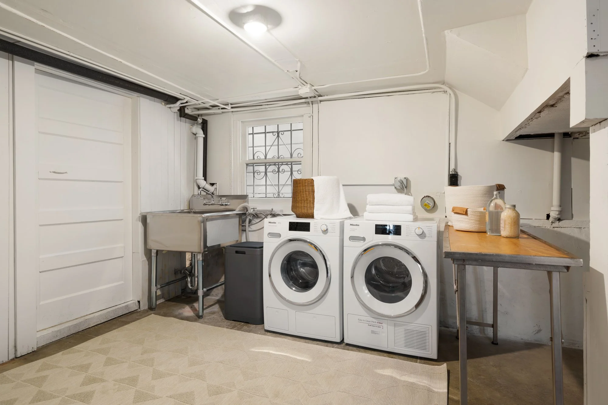 Laundry room with washing machine and dryer next to stainless steel sink, wooden folding table, and window above. Towels, a basket, and glass jars are on the table.