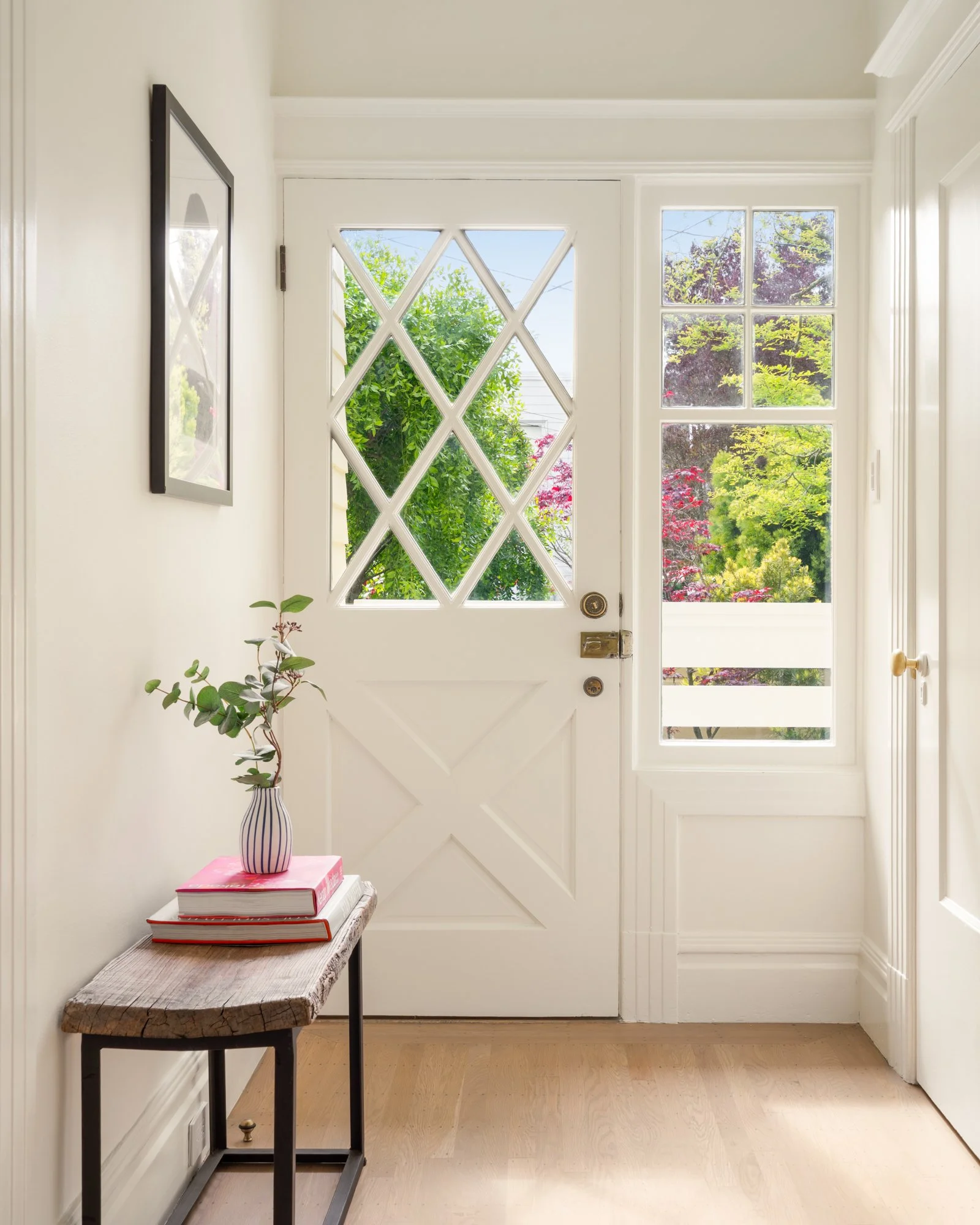 Bright entryway with a white door featuring diamond-shaped glass panes, adjacent window showing greenery outside, and a small wood table with a vase and stacked books.