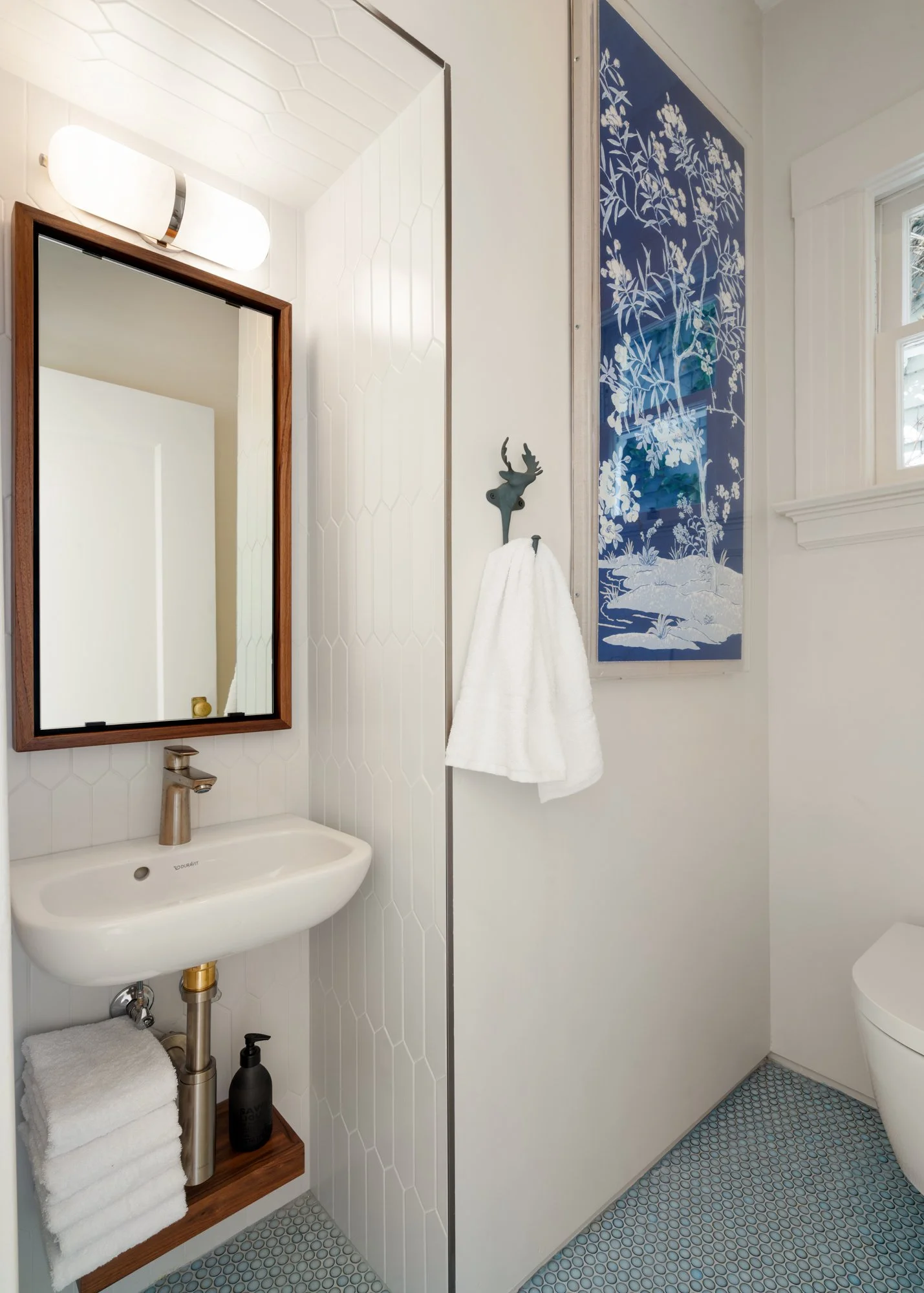 Modern bathroom with a small white sink, wooden framed mirror, wall-mounted light above, towel rack with white towel, art piece on the wall, and patterned floor tiles.