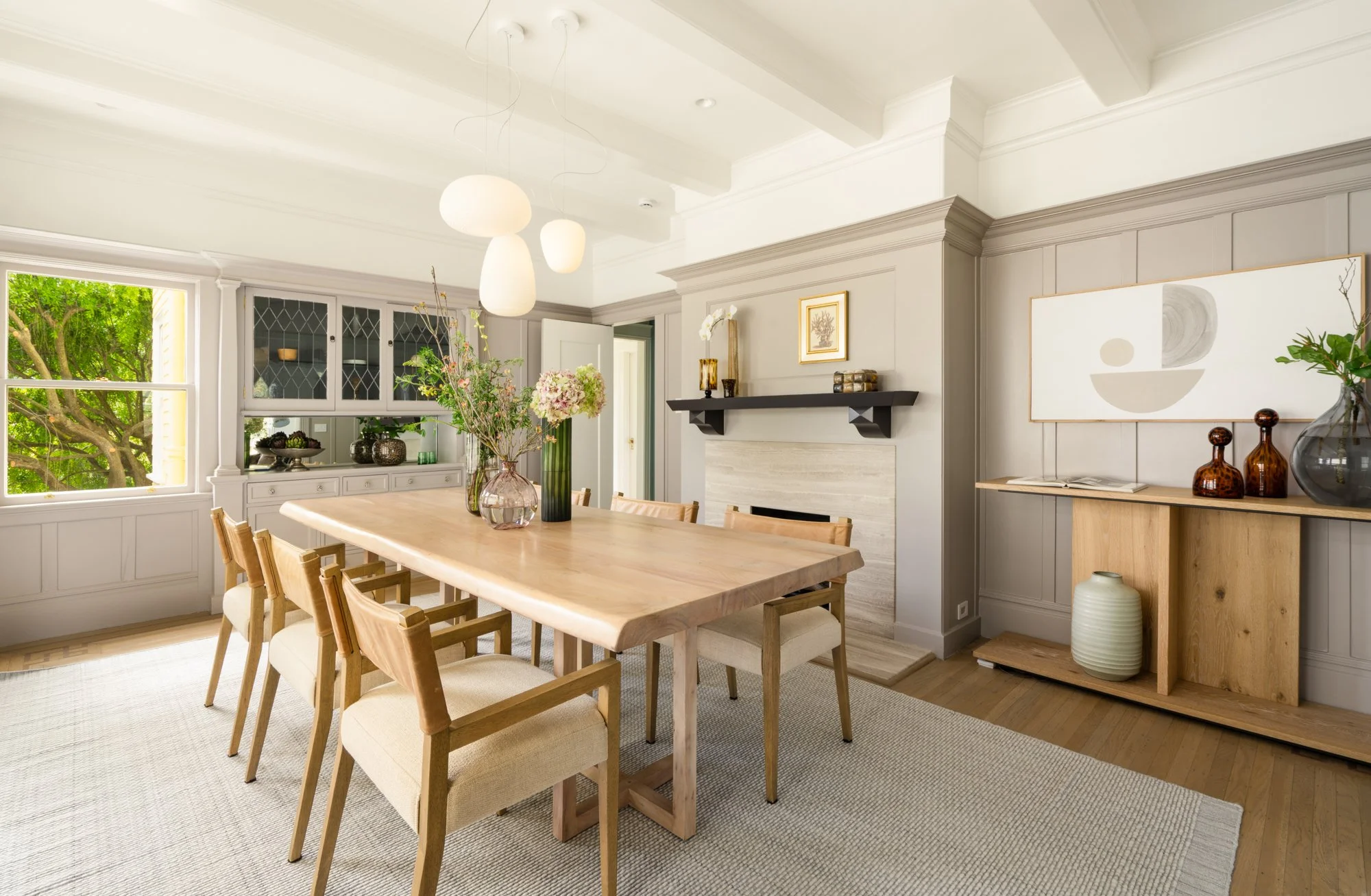 Modern dining room with wooden table and chairs, pendant lights, plants, large window, and abstract wall art.