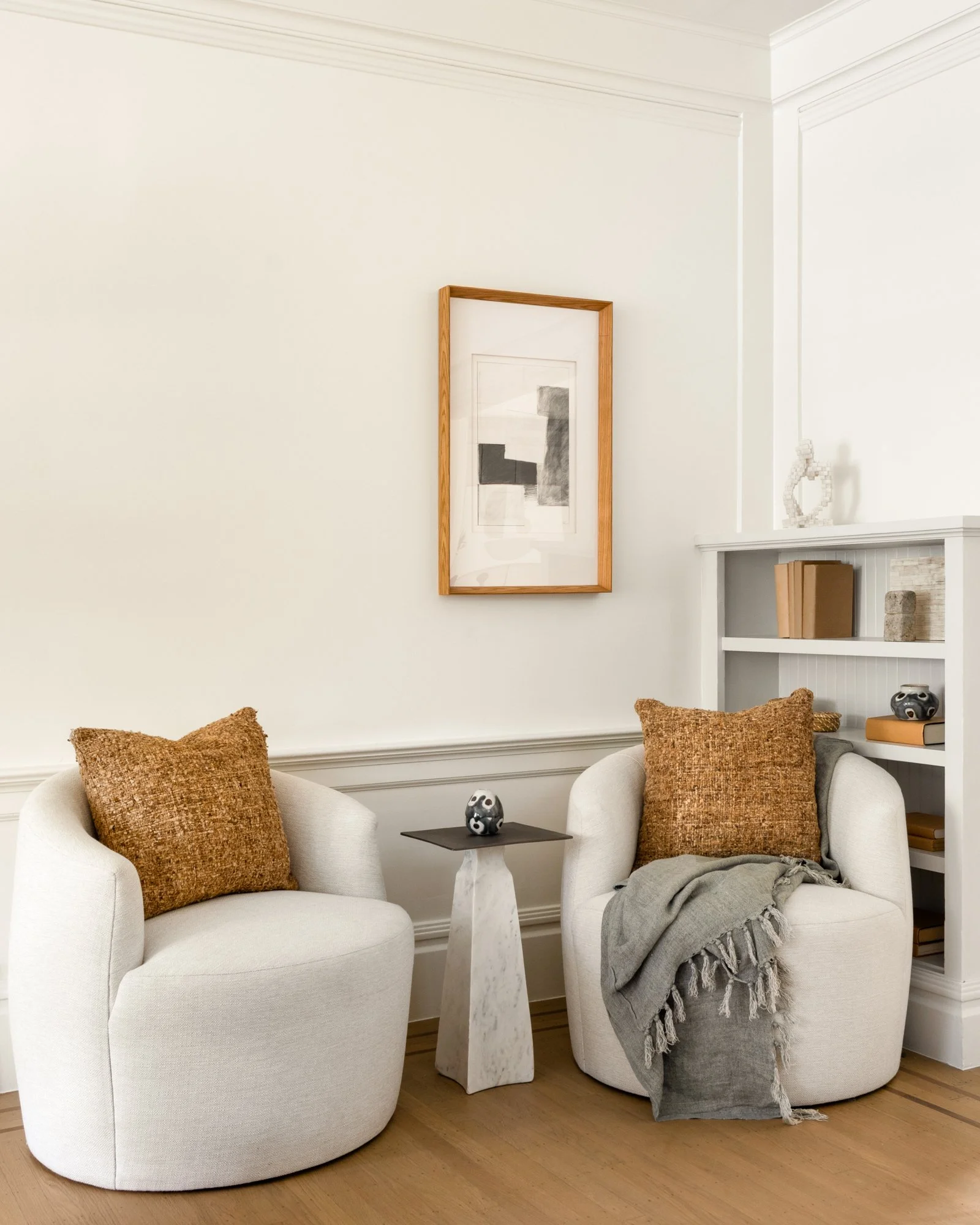 Cozy sitting area with two beige armchairs, brown textured cushions, a small marble side table, and abstract artwork on the wall. Shelves hold books and decorative items.