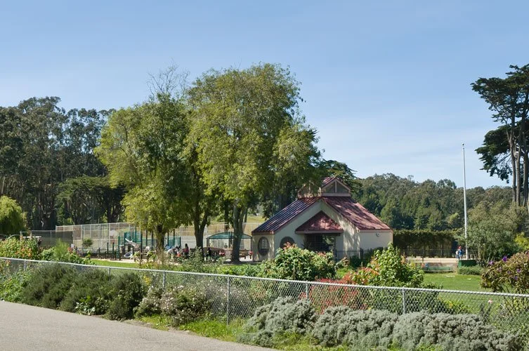 Outdoor park scene with trees, a small building, playground, and greenery.