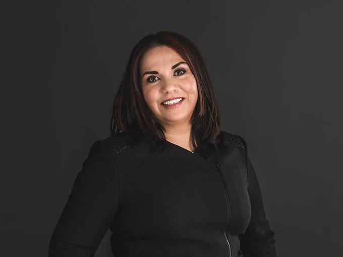A woman with dark hair smiling, wearing a black top, against a dark background.