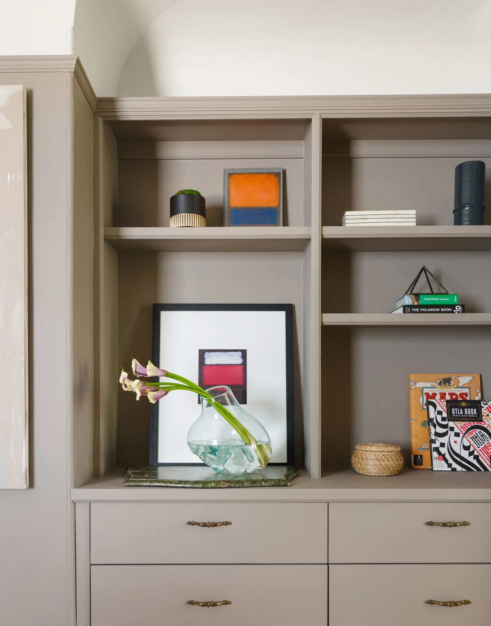 Interior design featuring a beige built-in shelving unit with decorative items including framed abstract art, a vase with flowers, books, and a small basket.