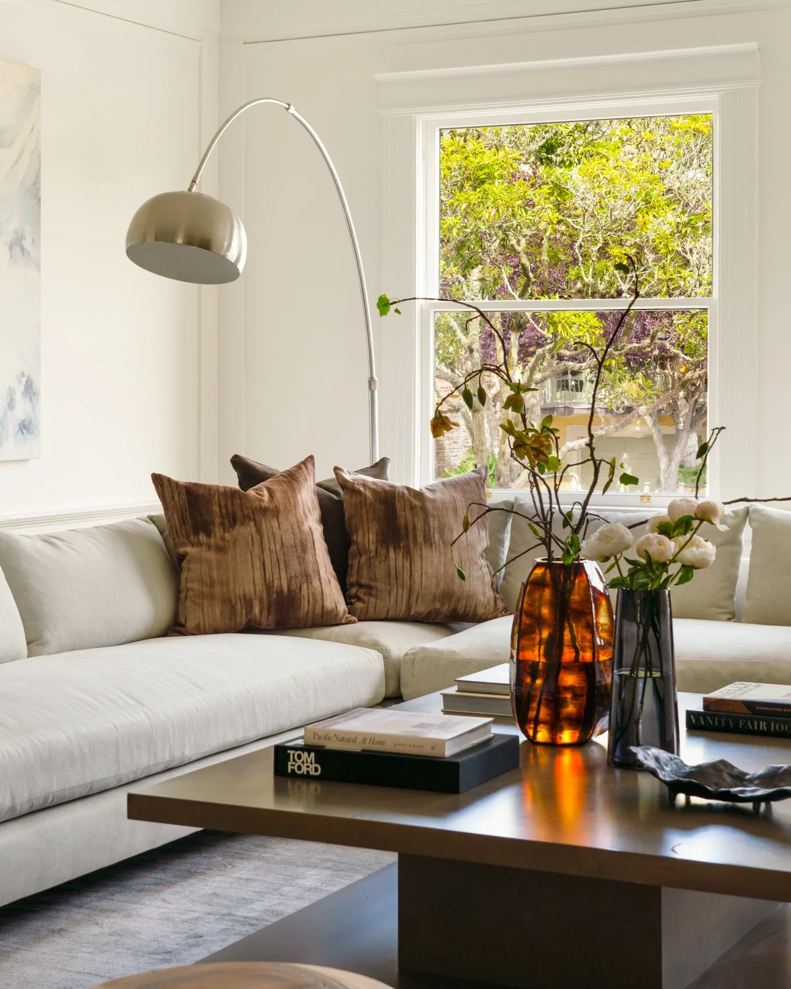Modern living room with beige sectional sofa, brown throw pillows, arched floor lamp, and a wooden coffee table adorned with books and decorative vases containing branches with white flowers. A large window in the background shows green trees outside