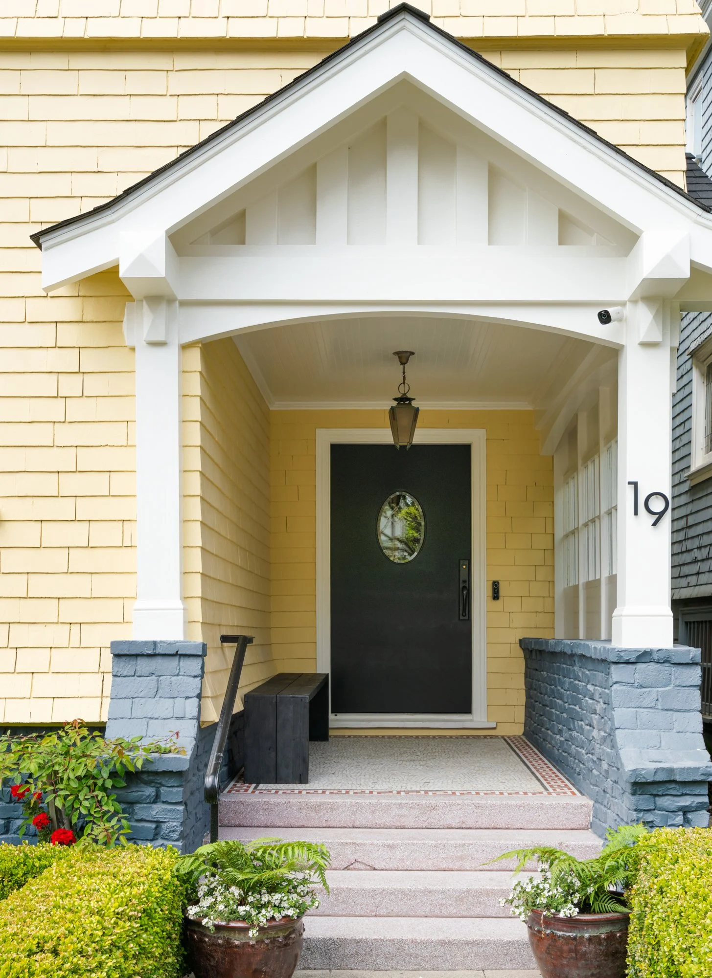Front entrance of a house with yellow siding, a black door with an oval window, white woodwork, a hanging light fixture, steps, potted plants, and shrubs.