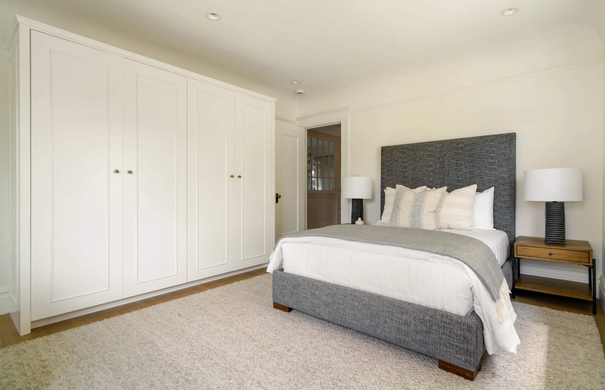 Modern bedroom with a gray upholstered bed, white pillows, wooden nightstands, and white closet doors.