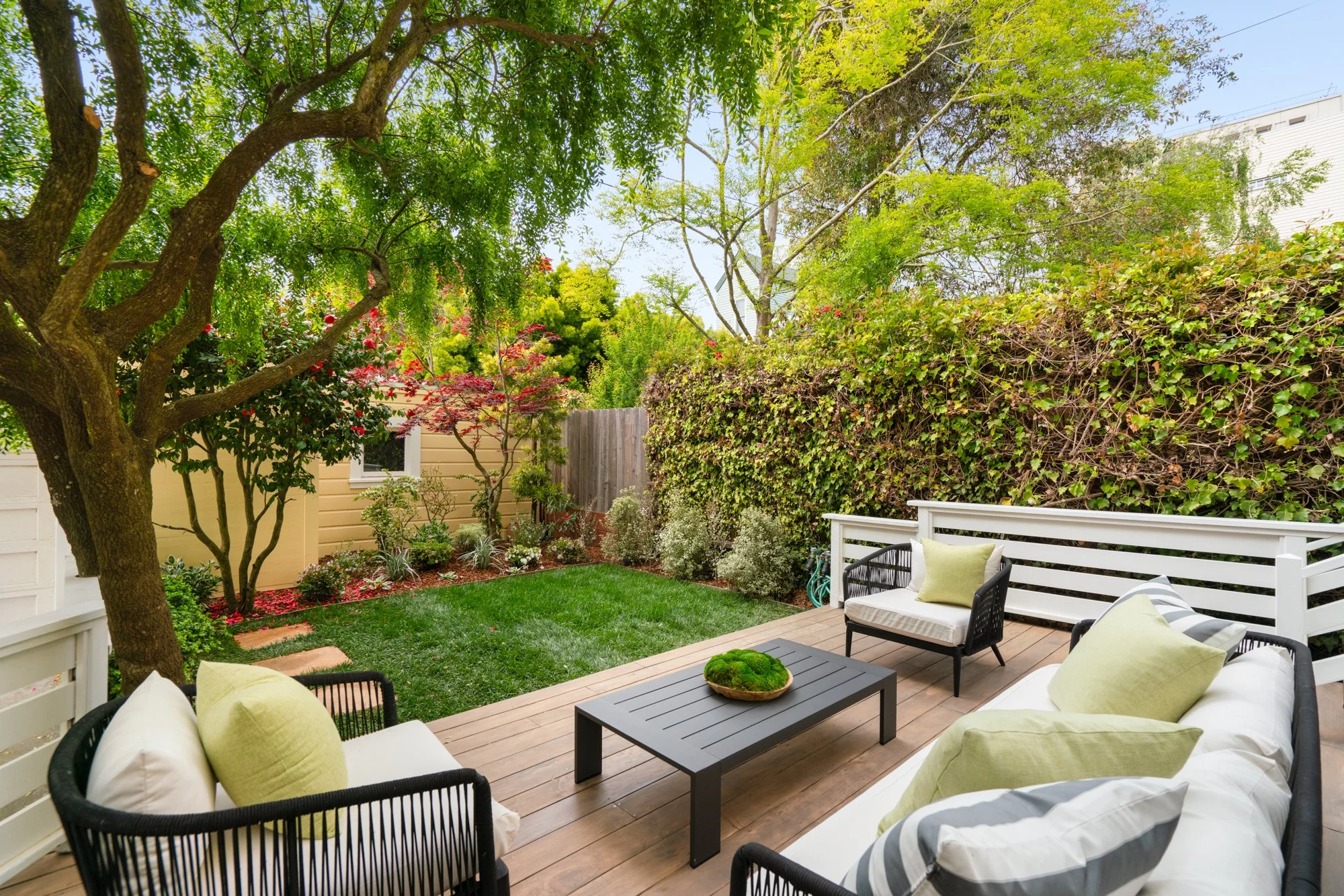 Outdoor patio with comfortable seating, green cushions, a black table, surrounded by lush garden with trees and shrubs on a sunny day.