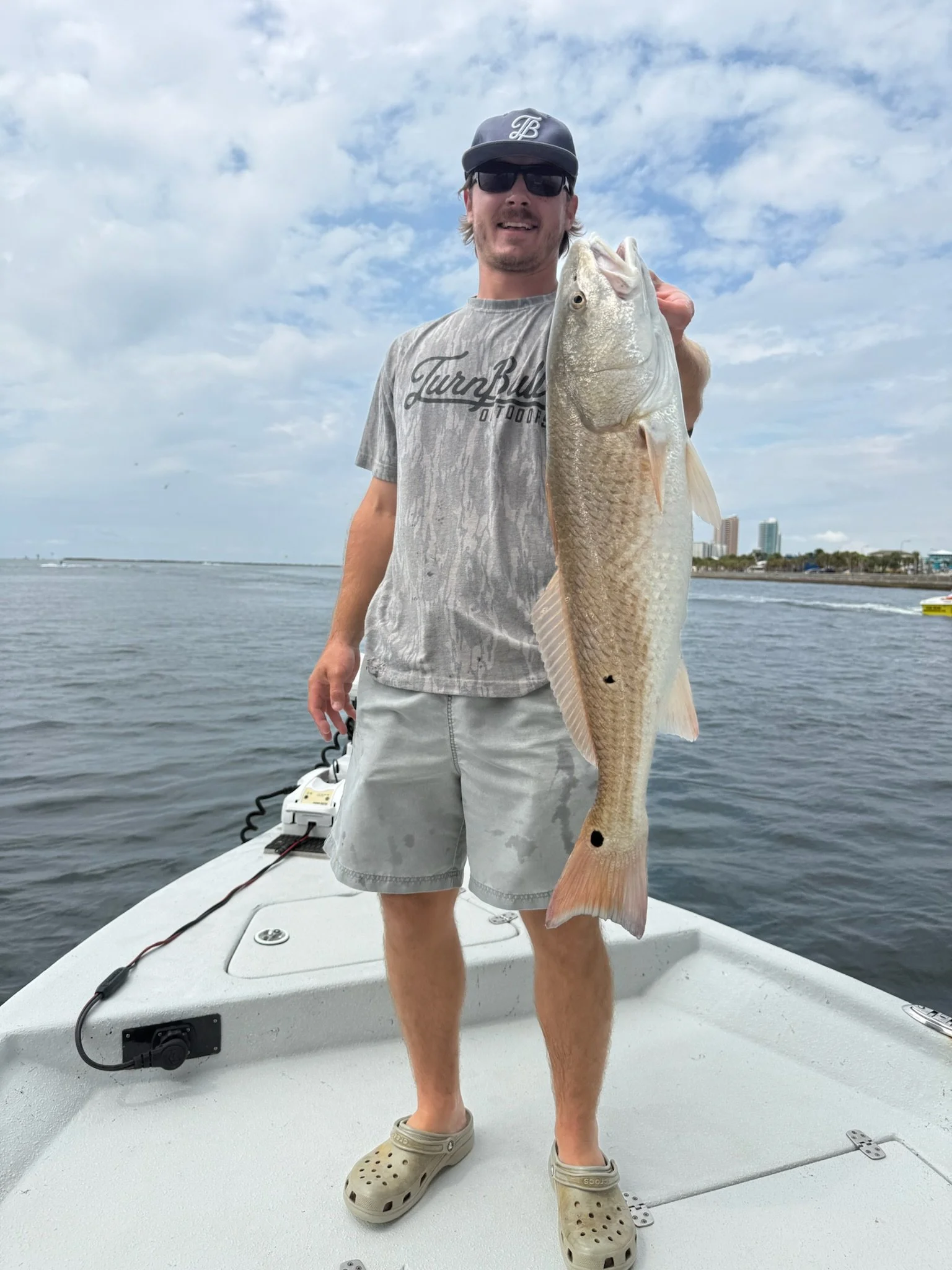 A man wearing a dark cap, sunglasses, and a light gray t-shirt holding a large fish on a boat with water and a city skyline in the background.