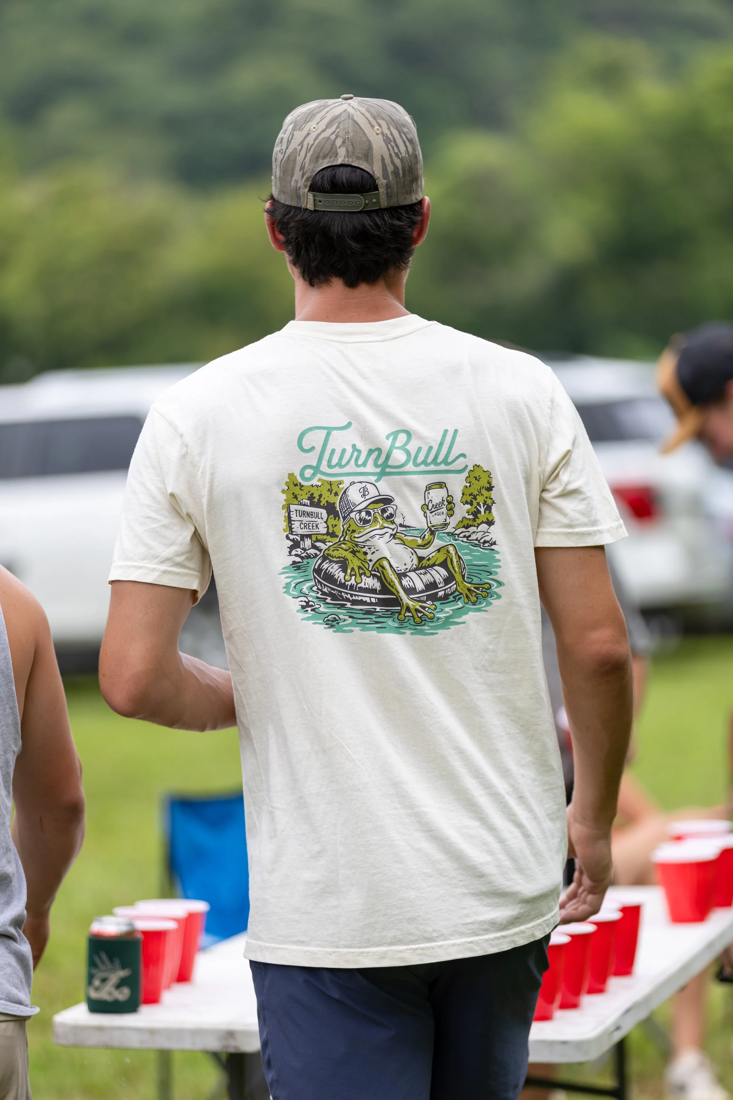 A man is seen from behind wearing a tan t-shirt with a cartoon frog graphic and the words "TurnBull" on the back, as he stands near a red cup relay game at an outdoor gathering.