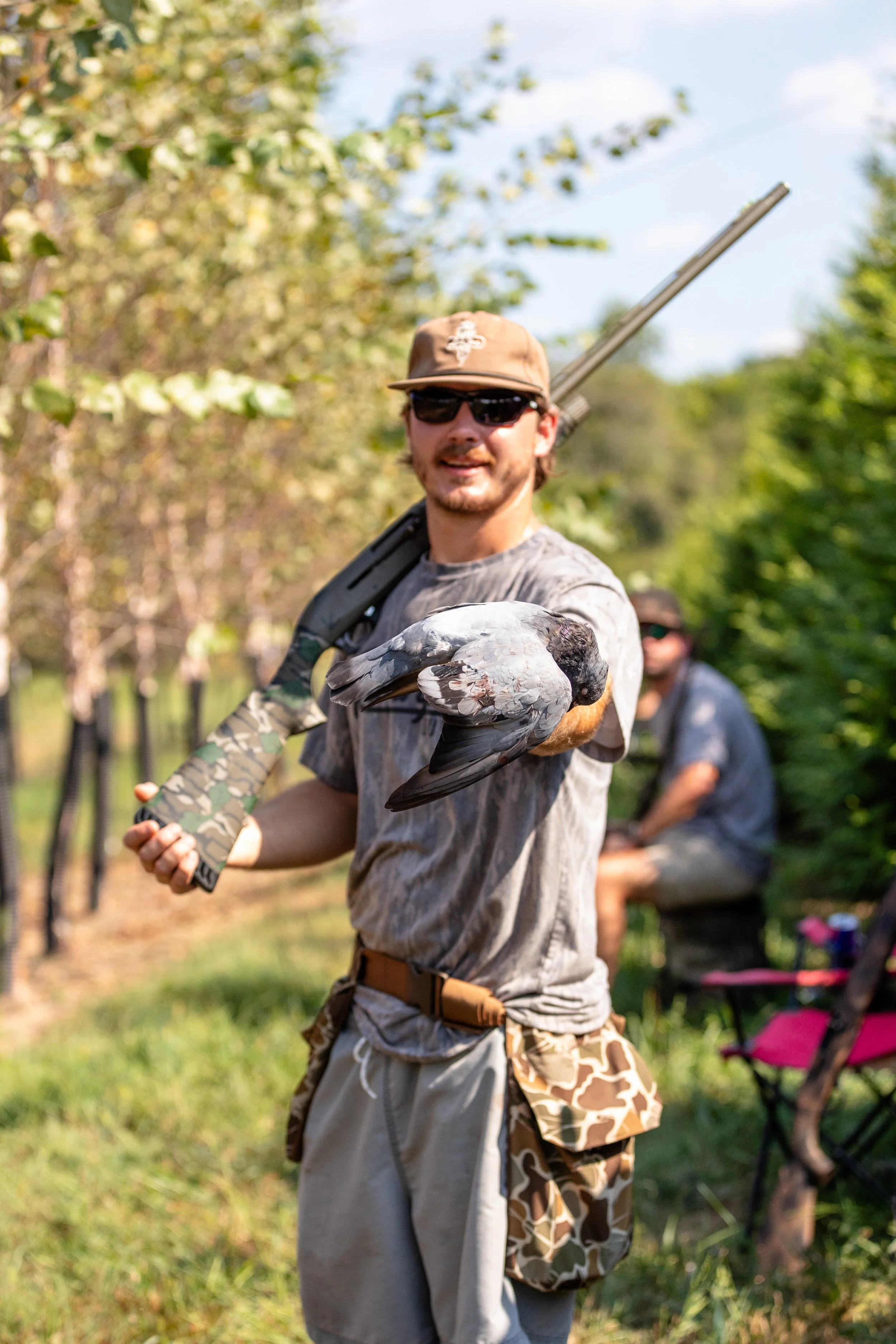 A man wearing a brown cap, sunglasses, and camouflage clothing holding a hawk on his gloved arm in a park setting with trees and a person sitting in the background.