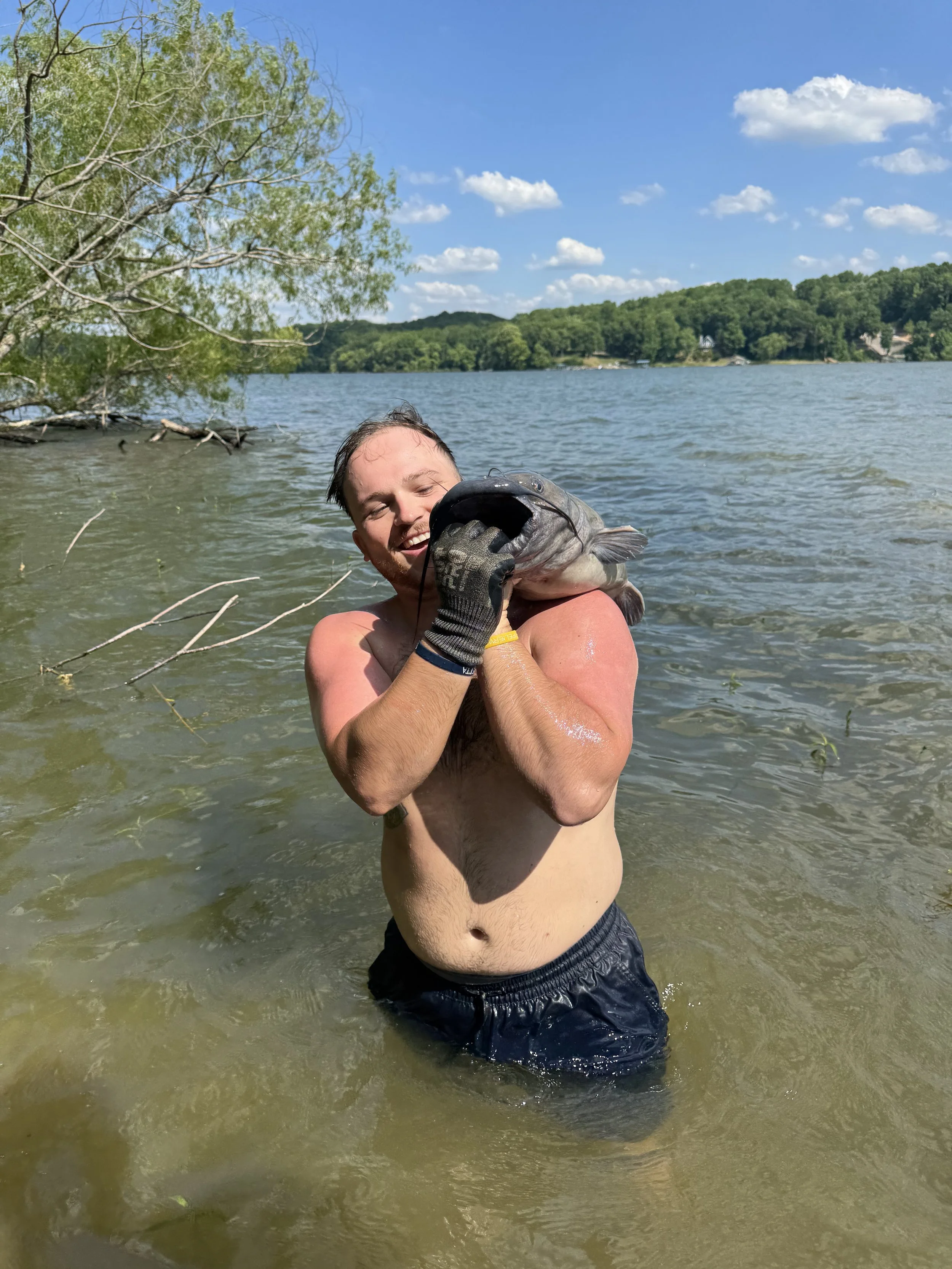 A man standing in a lake, holding a large fish with both hands while smiling. The man is shirtless and wearing black shorts, with green trees and a blue sky with clouds in the background.