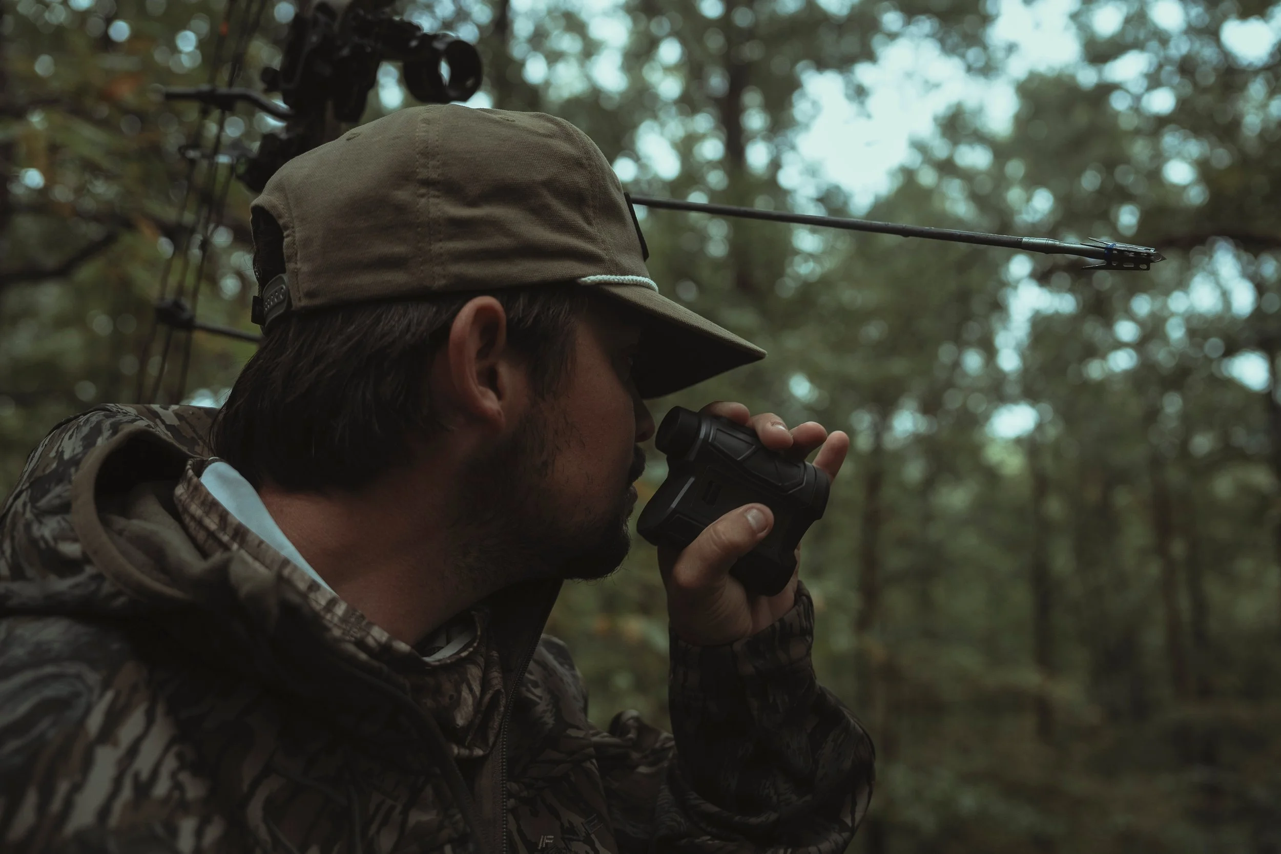 A person with a cap and camouflage jacket looking through binoculars in a forest with blurred trees in the background.