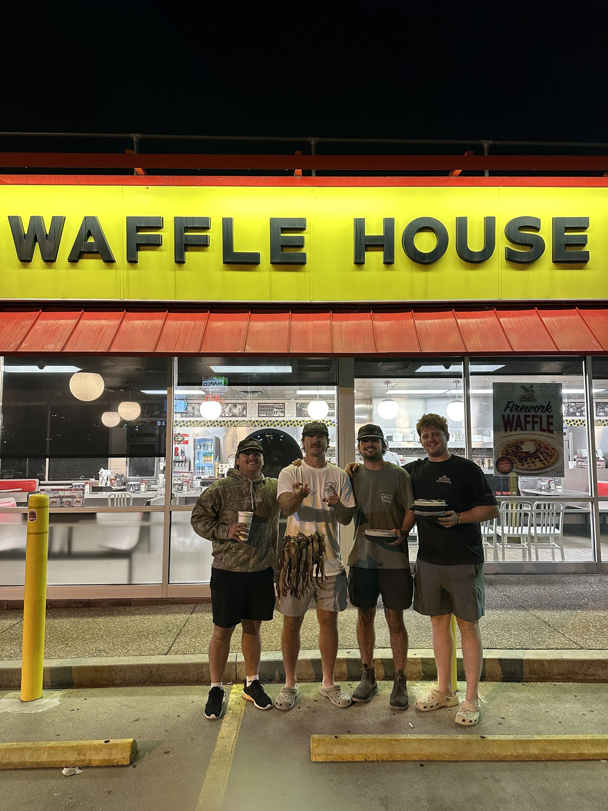 Four young men standing outside a Waffle House restaurant at night, smiling and posing for a photo, with some food and drinks in hand.