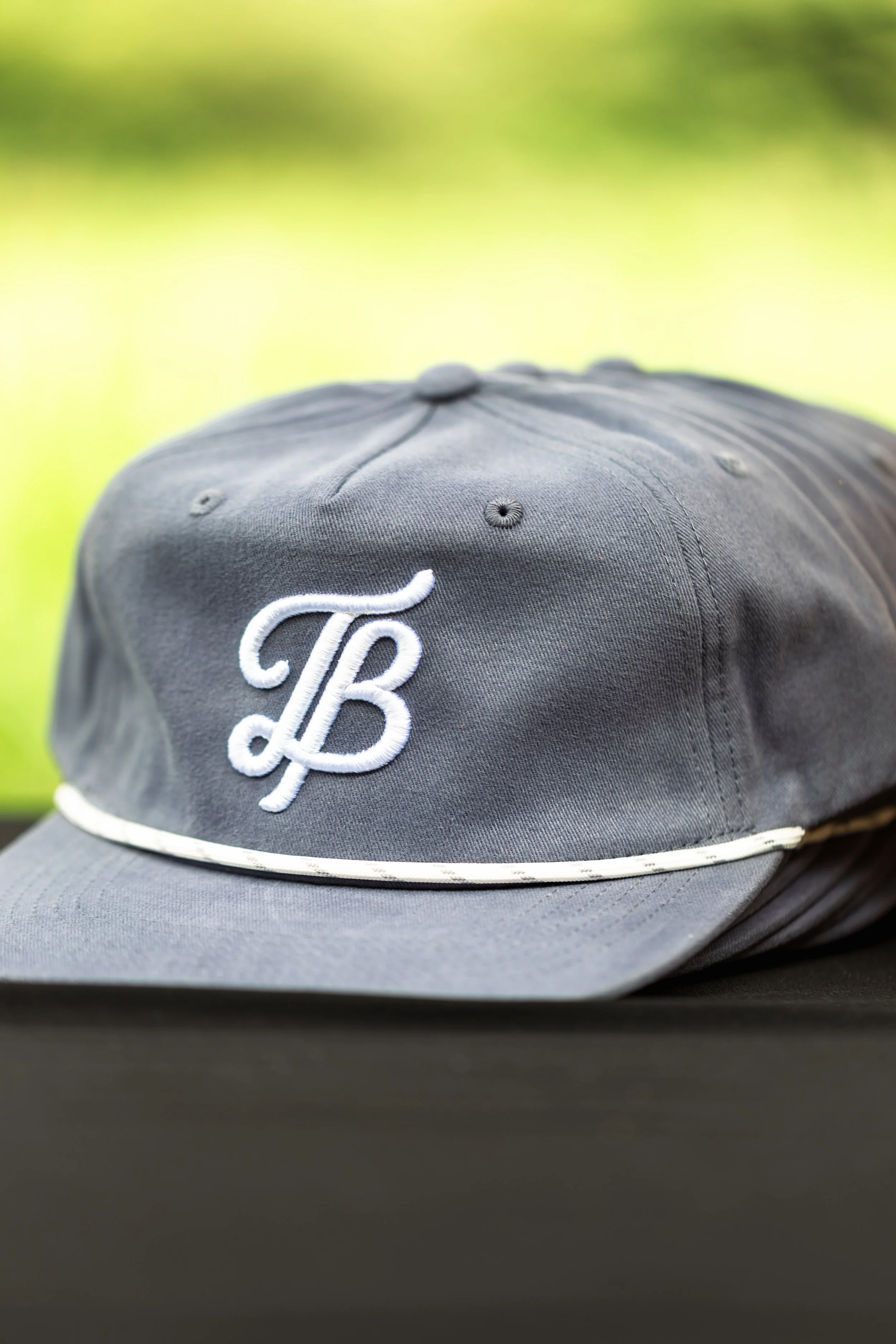 Close-up of a dark gray baseball cap with white embroidered initials 'TB' on the front and a white trim along the bill, set against a blurred outdoor background.