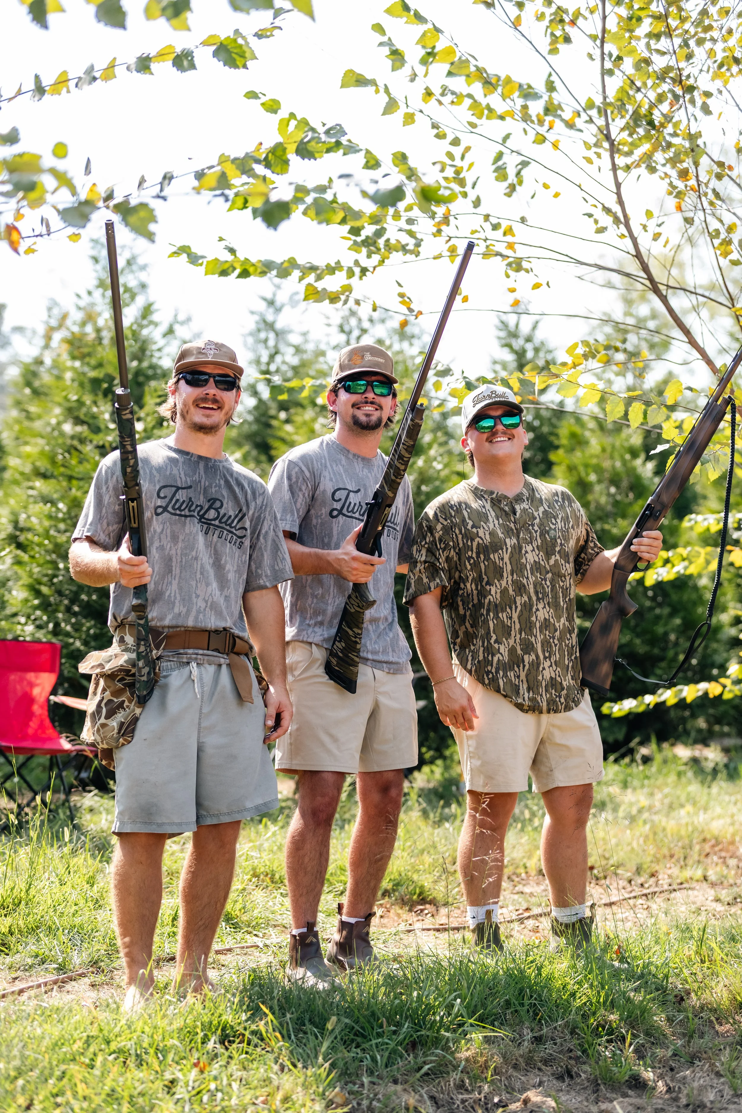 Three young men standing outdoors in a wooded area, holding rifles, smiling. They are wearing casual clothing, caps, and sunglasses, with green trees and a grassy field in the background.