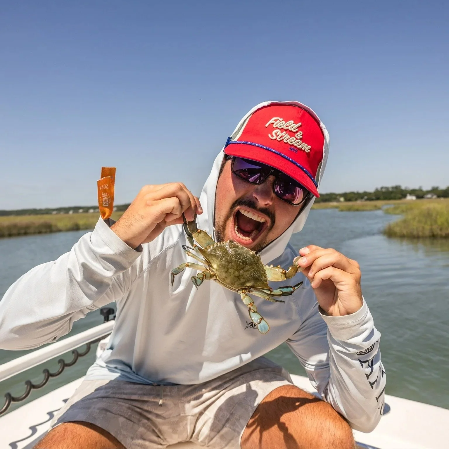 A man wearing sunglasses, a white hoodie, and a red cap holding a crab on a boat in a marshy area.