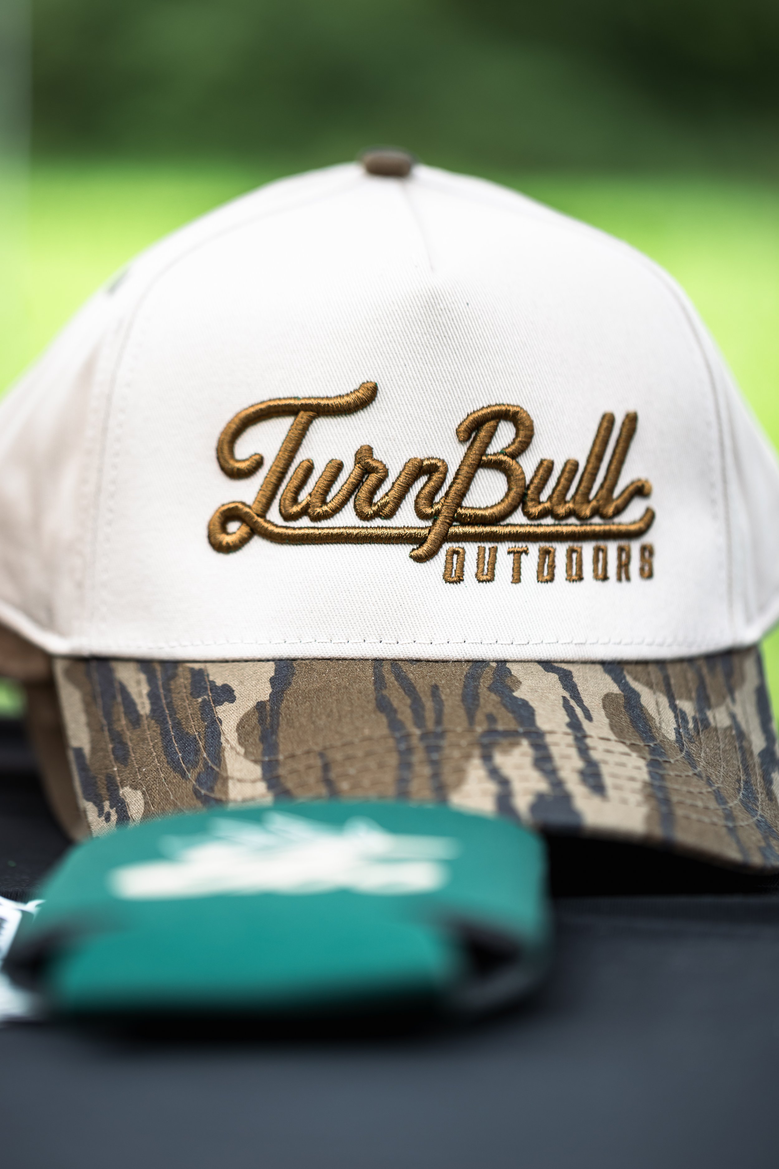 Close-up of a white baseball cap with brown embroidery that reads "LureBully Outdoors". The cap is partially pulled down over a camouflage-patterned item.
