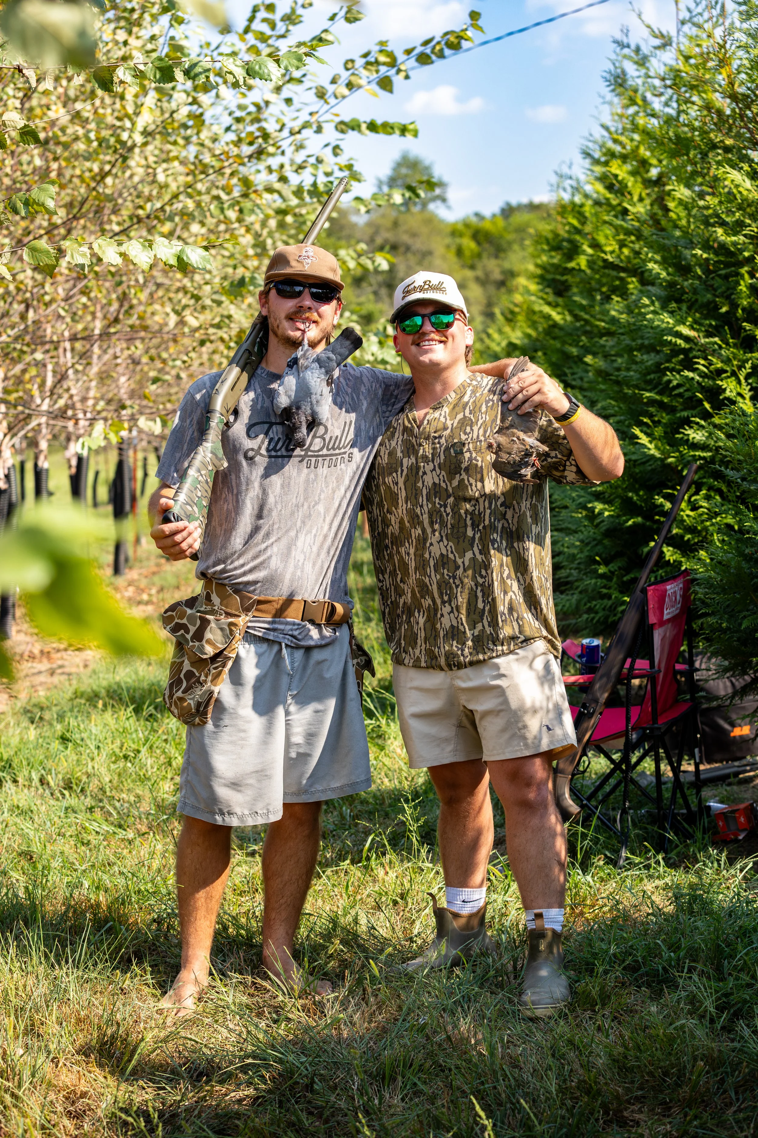 Two men standing outdoors in a green, wooded area, smiling and posing with a bird they caught. One man is holding a shotgun, the other has his arm around him, and they are dressed in outdoor, camouflage, and casual clothing.