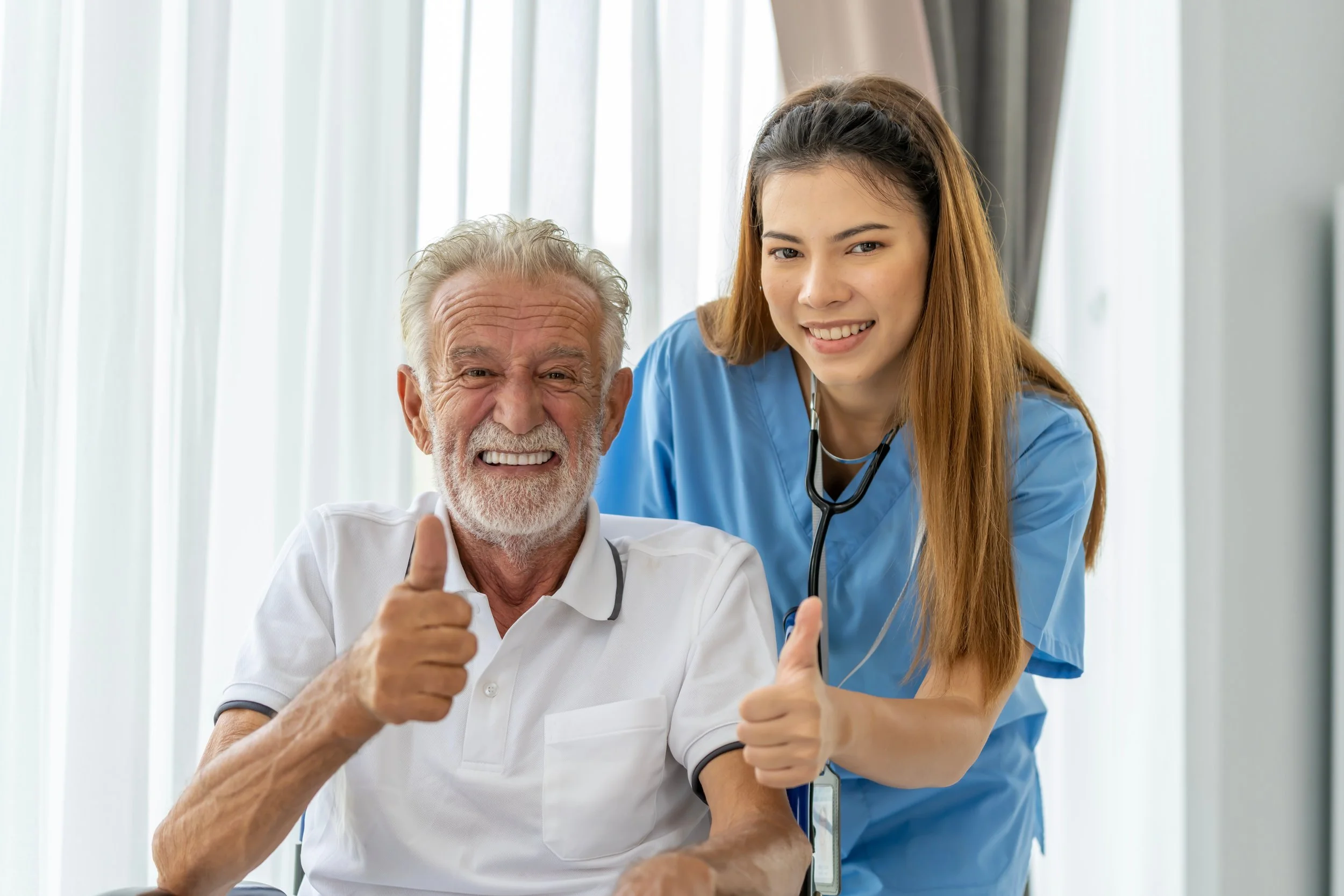 Elderly man with white hair and beard giving a thumbs up, sitting in a medical setting with a young female healthcare professional in blue scrubs standing beside him, also giving a thumbs up.