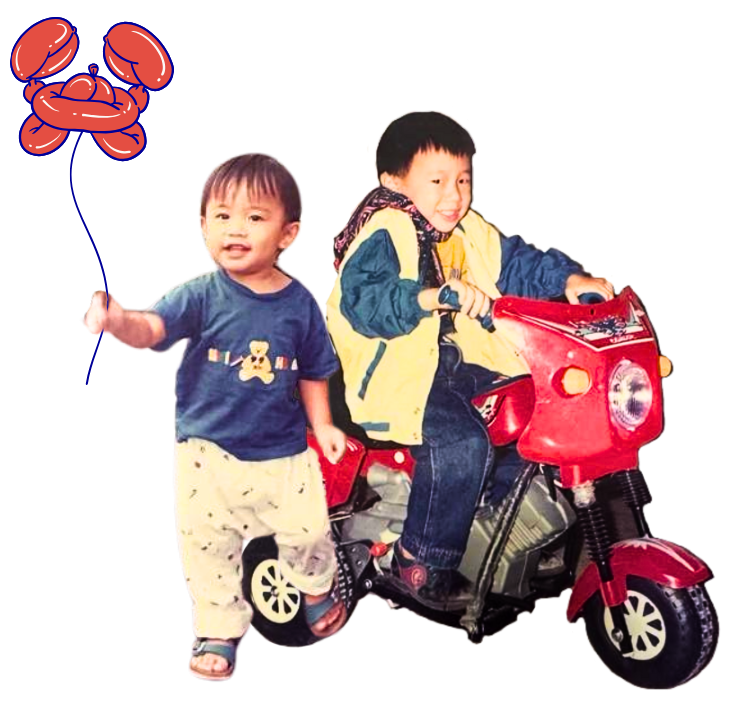 Two young boys playing together, one riding a red toy motorcycle and the other standing next to it.