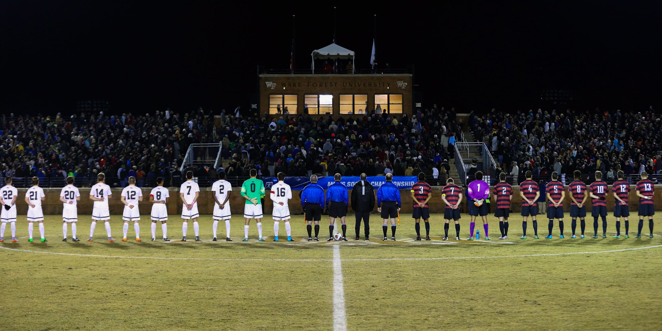 Soccer players standing in a line on the field before a match at Wake Forest University, with spectators in the stands behind them at night.